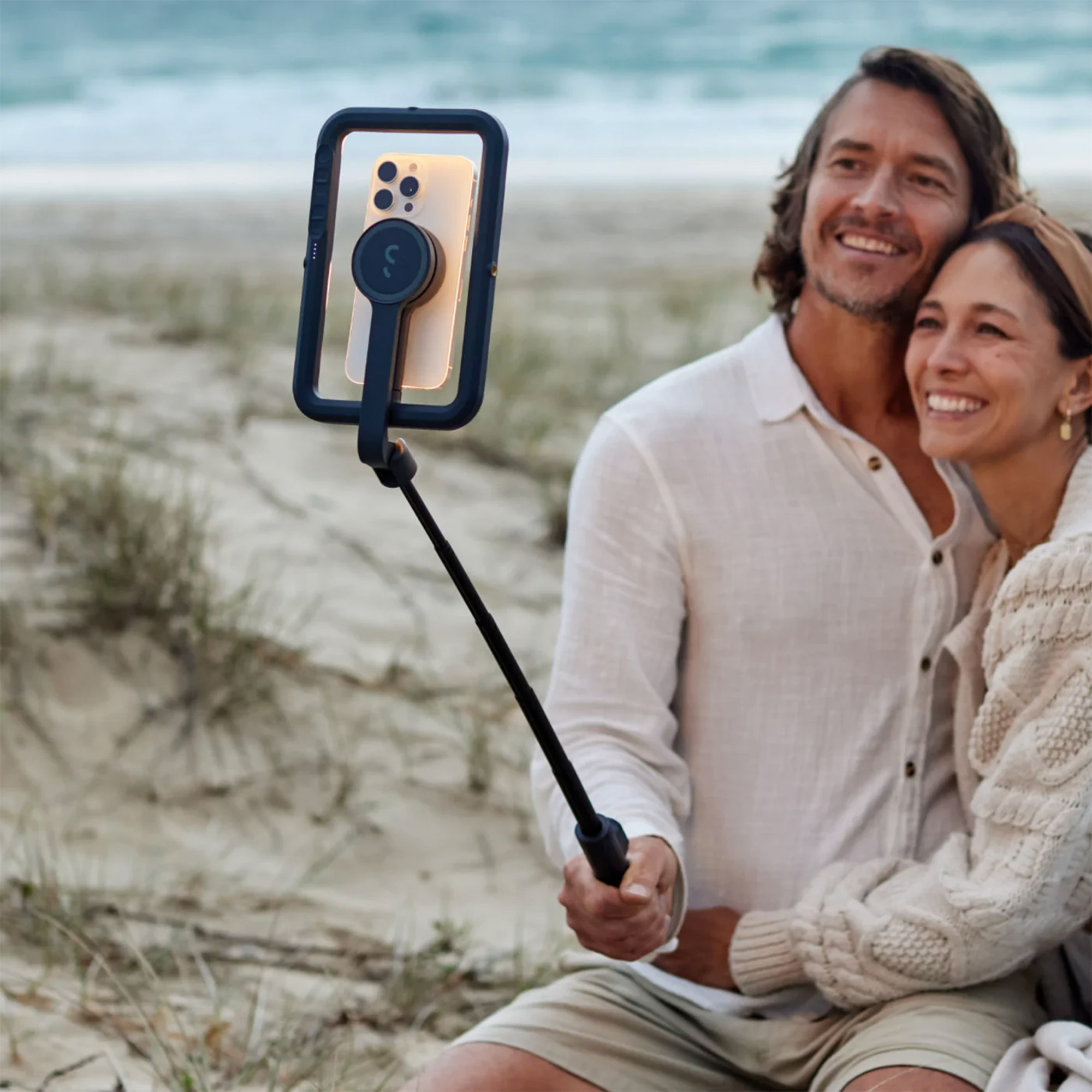 Sahilde telefonla selfie çeken bir çift. Gülümseyerek selfie çubuğunu tutuyorlar. Arka planda kum ve deniz var.