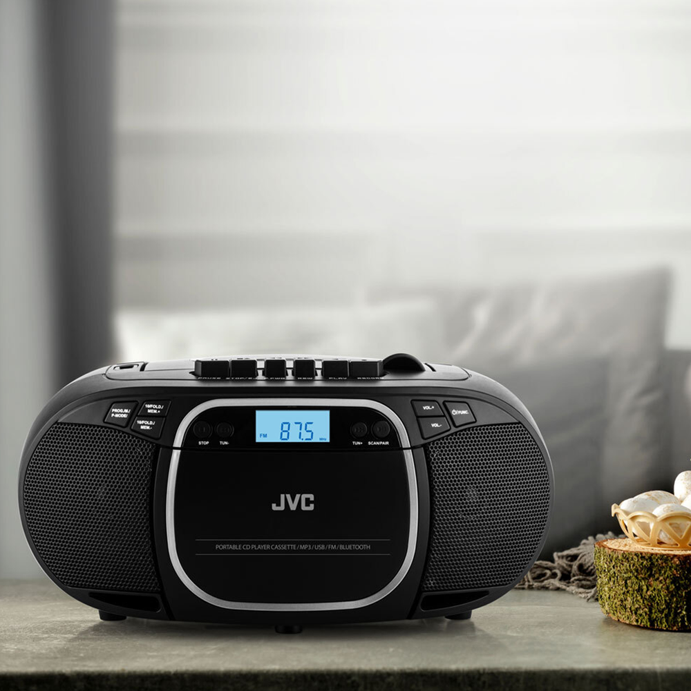 Czarny przenośny odtwarzacz CD JVC z radiem FM na stole. Wyświetlacz pokazuje 87,5 MHz. Tło: rozmyty pokój z poduszkami.