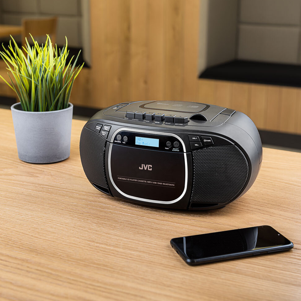 Czarny boombox JVC i smartfon na drewnianym stole. Doniczkowa roślina po lewej stronie. Tło rozmyte.