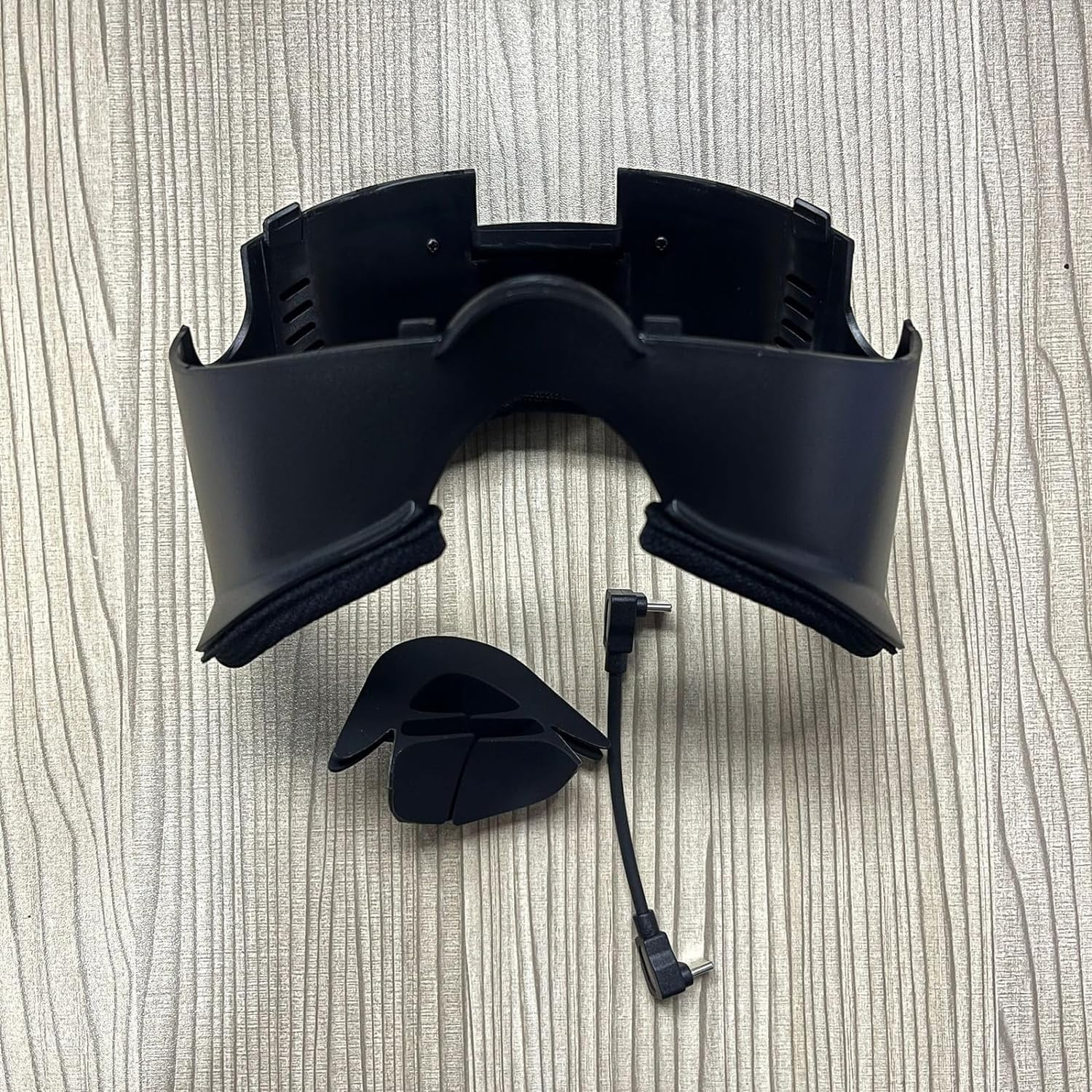 Ein schwarzes VR-Headset, ein Gesichts-Polster und ein Kabel mit abgewinkelten Anschlüssen liegen auf Holz.