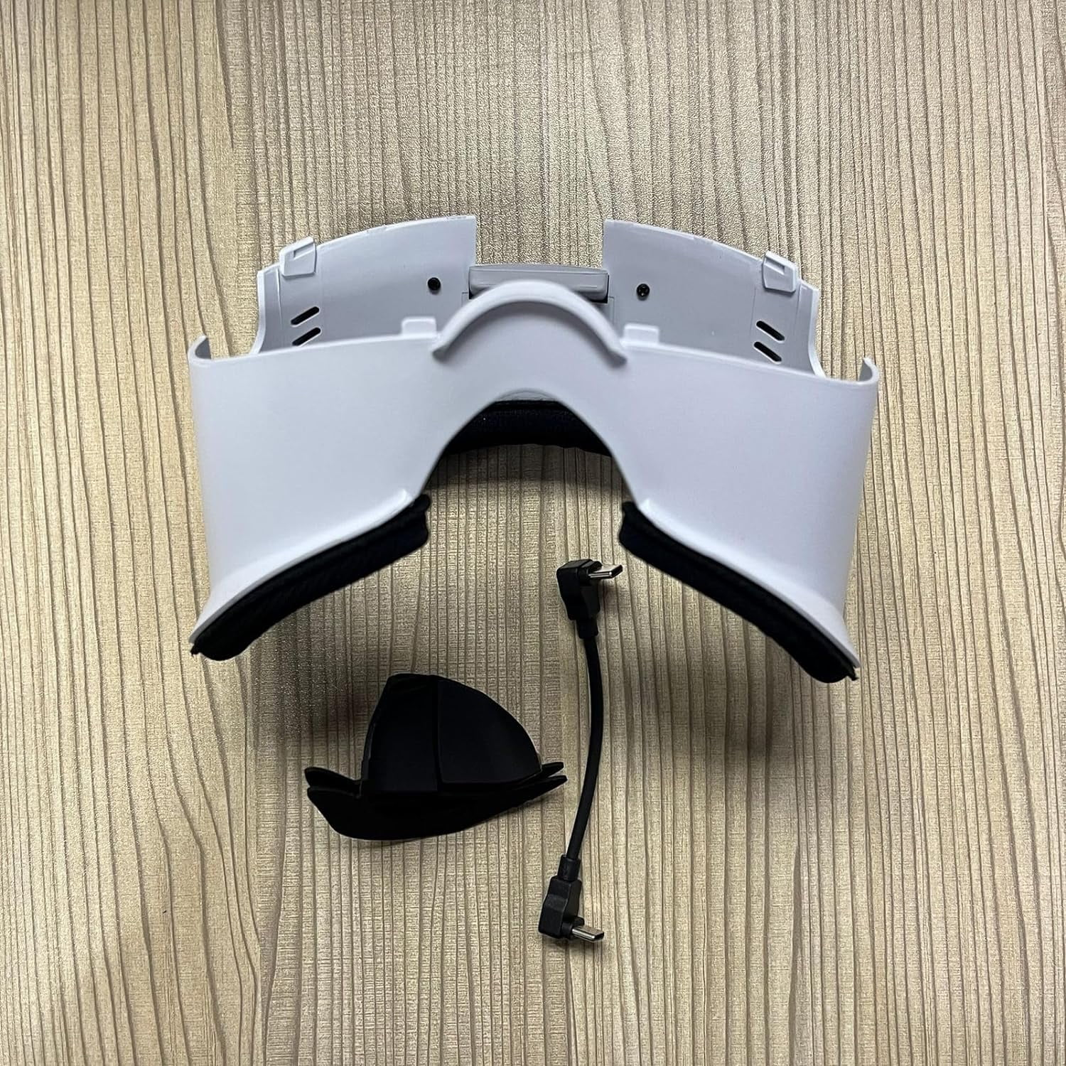 Ein weißes VR-Headset, ein schwarzes Kissen und ein schwarzes Kabel mit Stecker, alles auf einer Holzoberfläche.