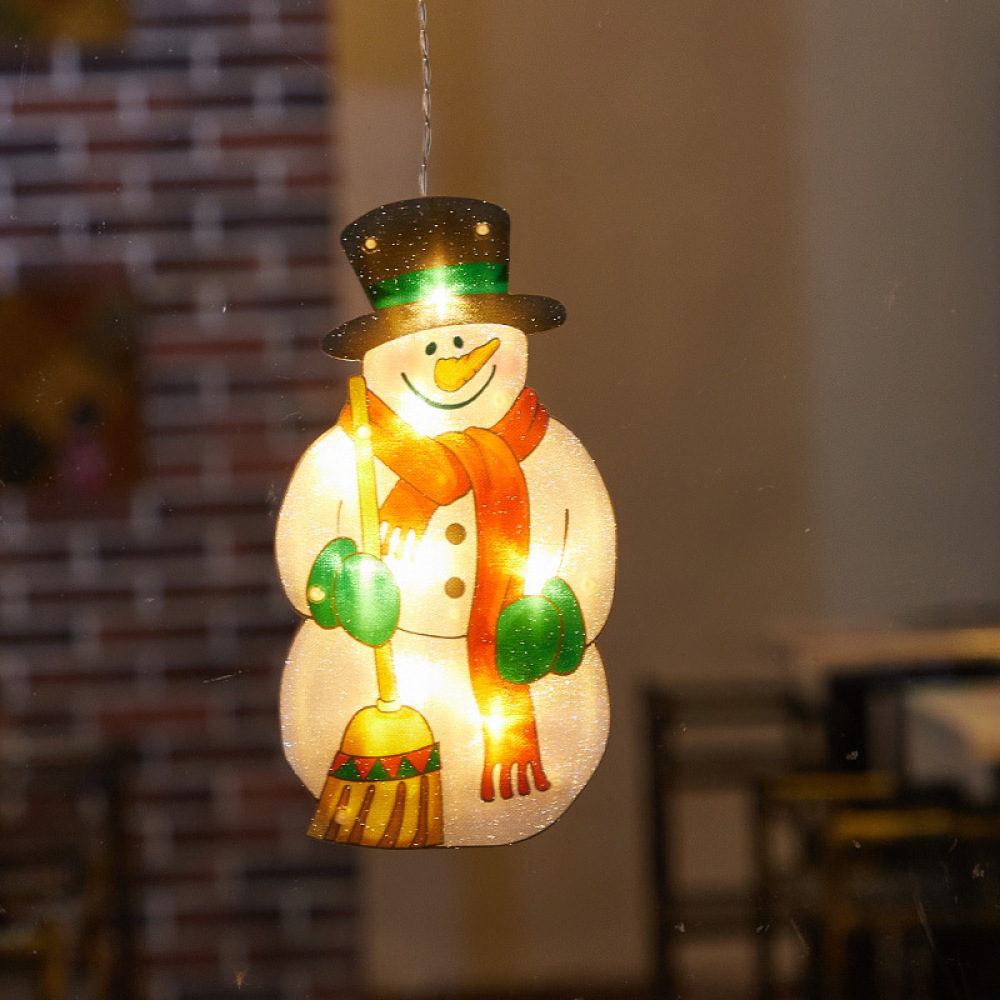 Eine Schneemann-Dekoration mit schwarzem Hut, orangefarbenem Schal, grünen Handschuhen und einem Besen. Es ist mit Lichtern beleuchtet und hängt.