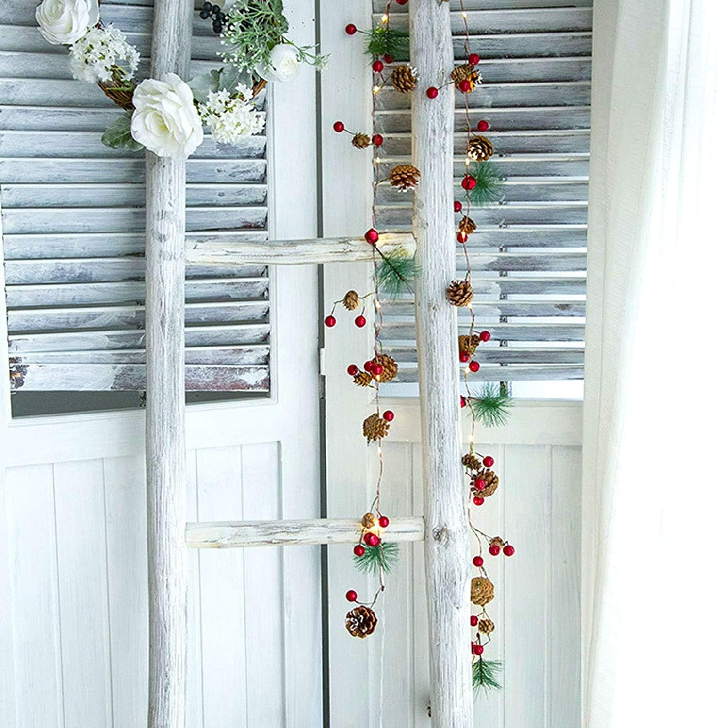 Eine weiße Leiter, verziert mit Tannenzapfen, roten Beeren und einem Blumenkranz. Vor einem weißen Fensterladen.
