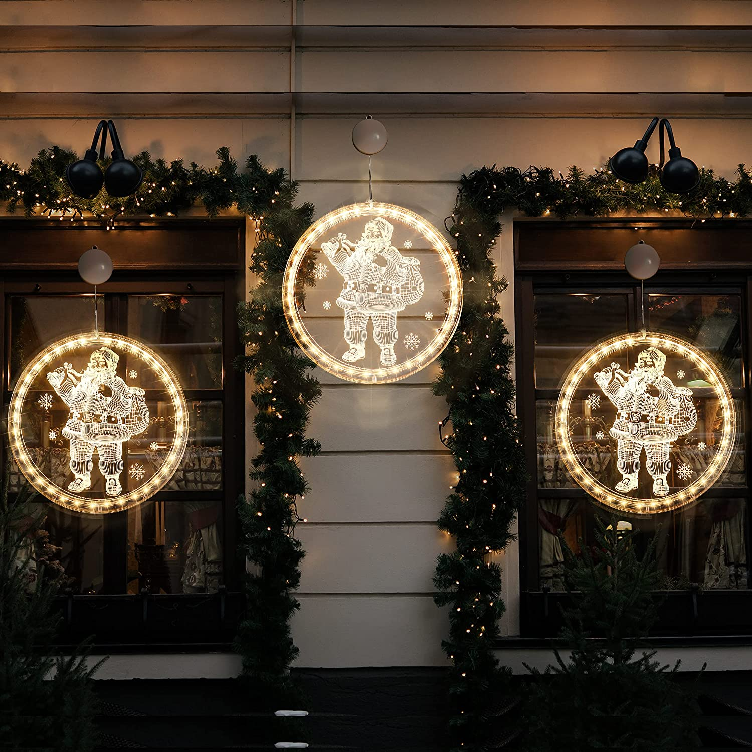 Drei beleuchtete Weihnachtsmann-Dekorationen hängen draußen an einem Gebäude. Jedes ist in einem Kreis, mit Schneeflocken. Grüner Girlandenschmuck.