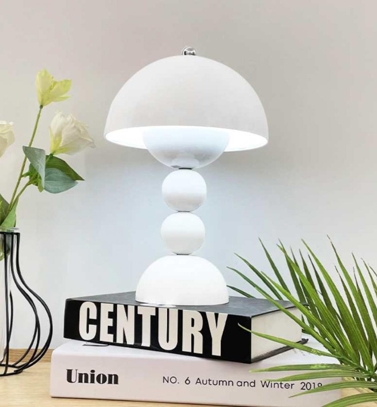 Eine weiße Lampe steht auf Büchern mit den Aufschriften Century und Union. Blumen und eine Pflanze befinden sich auf beiden Seiten der Lampe.