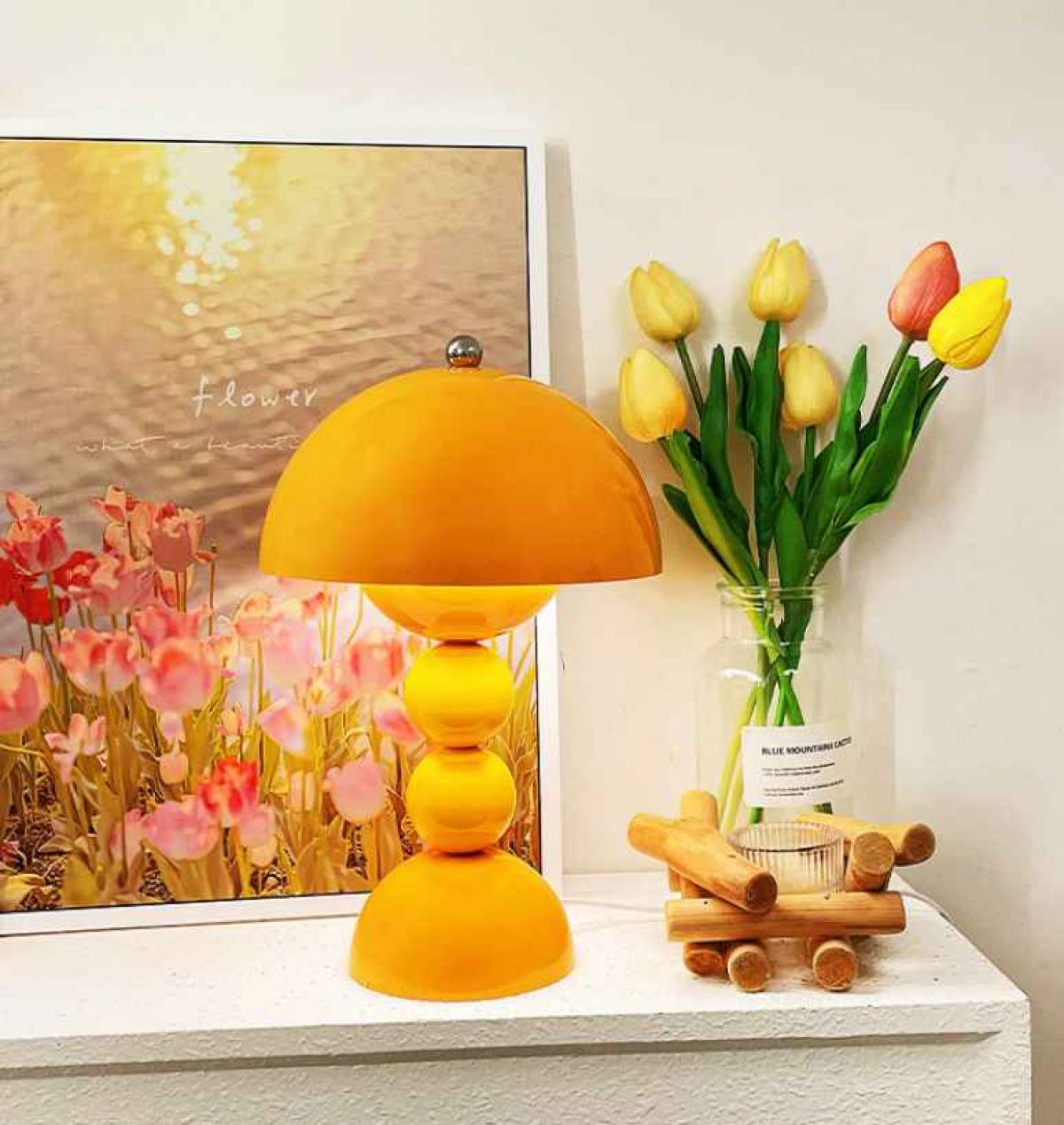 Eine orangefarbene Lampe, Tulpen und ein Kerzenhalter stehen auf einer weißen Oberfläche. Im Hintergrund befindet sich ein gerahmtes Bild mit Blumen.