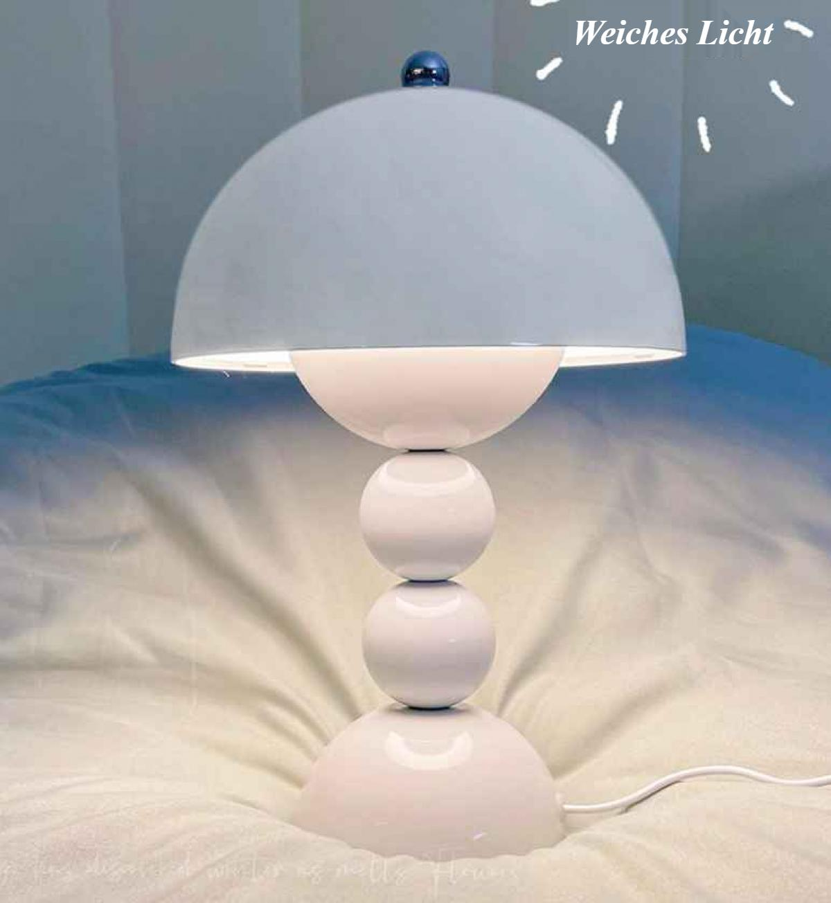 Eine weiße Lampe mit halbkugelförmigem Schirm steht auf einem Bett. Sie hat drei gestapelte Kugeln als Basis. Eine blaue Kugel krönt den Schirm.