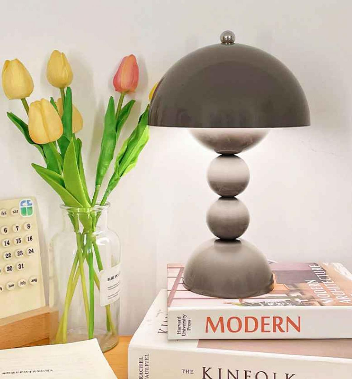 Eine moderne Lampe steht auf Büchern. Links steht eine Vase mit Tulpen. Ein Kalender ist ebenfalls vorhanden.