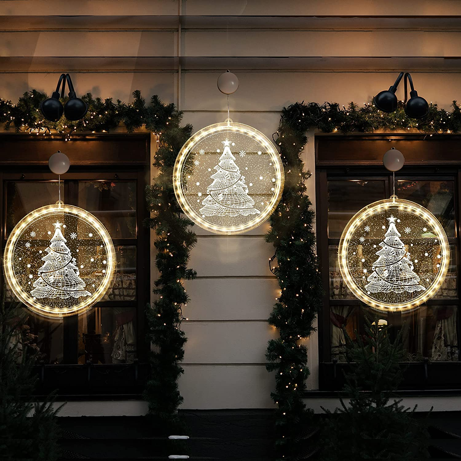 Drei beleuchtete Weihnachtsbaumdekorationen hängen draußen. Jede ist eine weiße Baumform in einem leuchtenden Kreis. Grünzeug um die Fenster.
