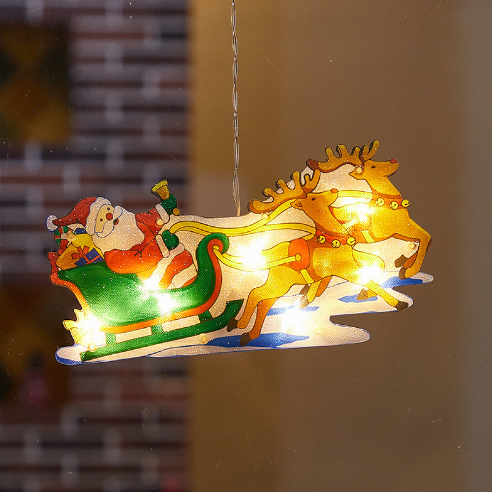 Eine Weihnachtsdekoration zeigt den Weihnachtsmann in einem von Rentieren gezogenen Schlitten. Die Dekoration ist beleuchtet und hängt.