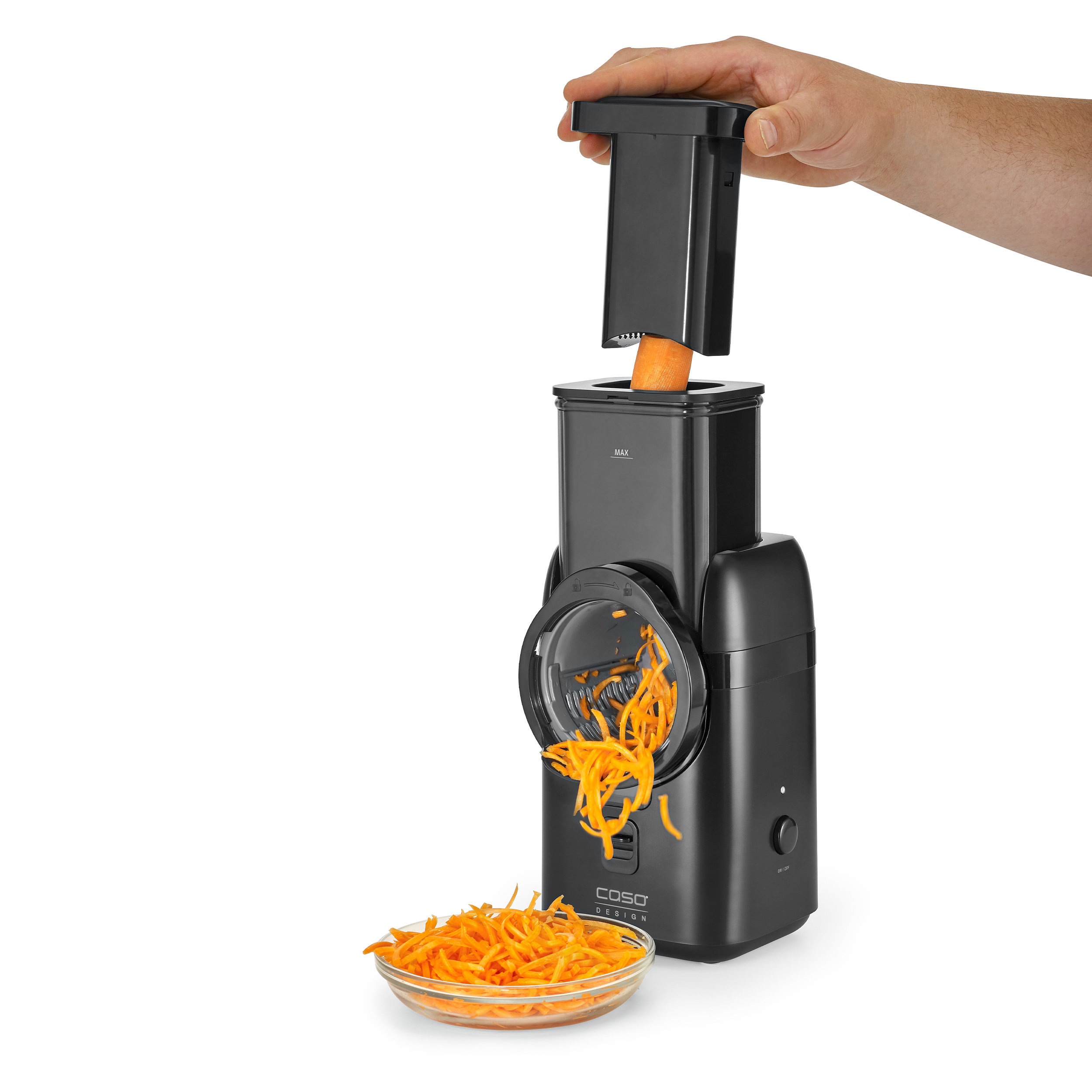 Una mano inserisce una carota in un robot da cucina nero. Carote grattugiate in una ciotola sotto. Sfondo bianco.