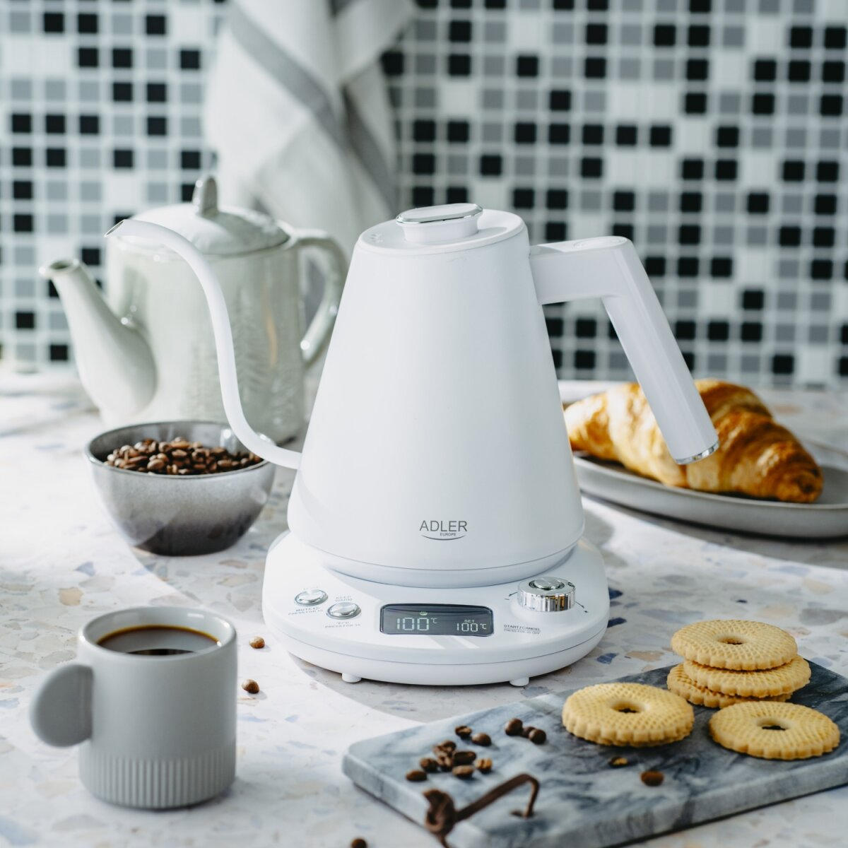 Weißer Wasserkocher mit Croissant, Keksen, Kaffee und Bohnen in einer Küche. Eine Teekanne ist ebenfalls zu sehen.