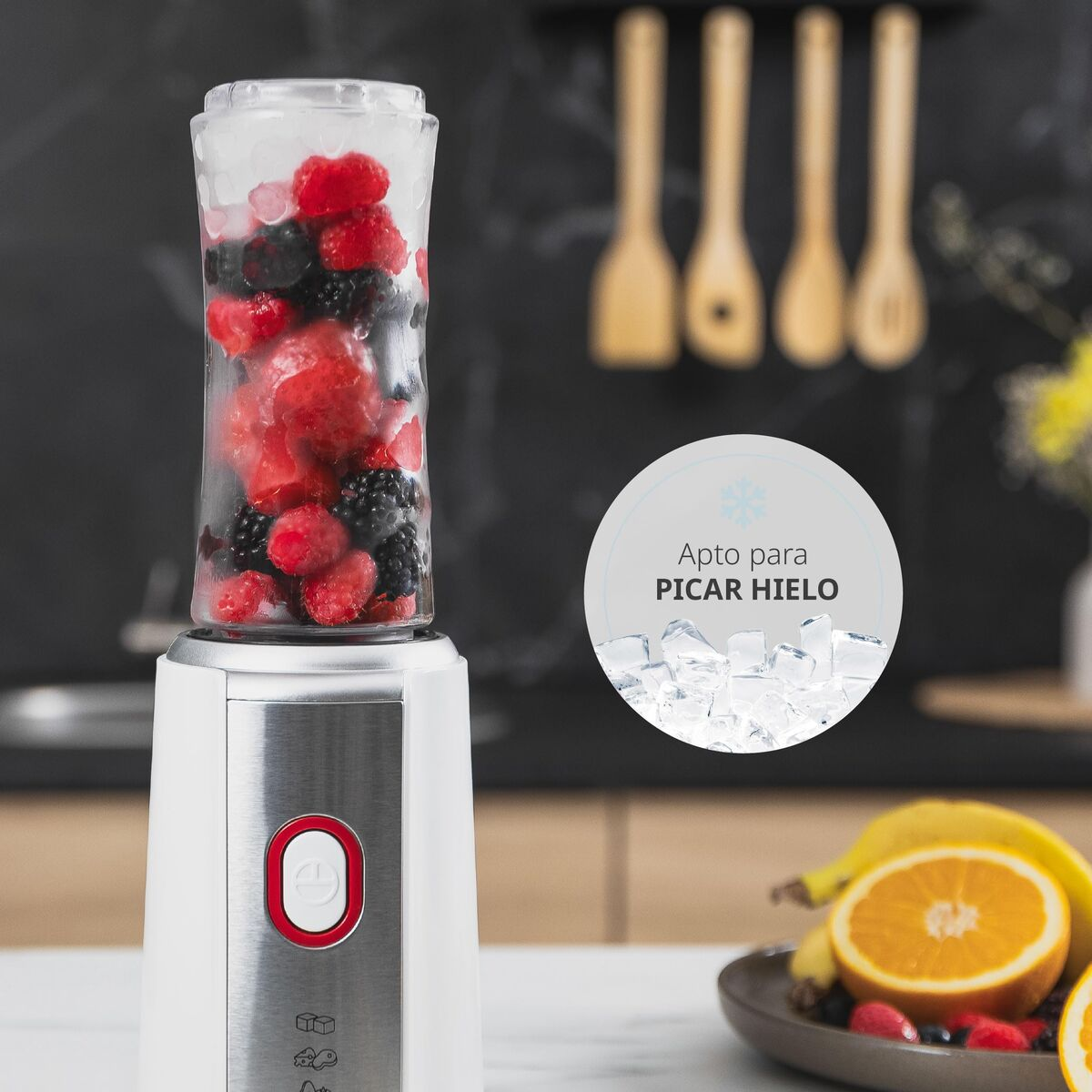 Ein Mixer mit gemischten Beeren und Eis. Rechts ein Teller mit Früchten und einer halbierten Orange. Im Hintergrund hängen Holzutensilien.