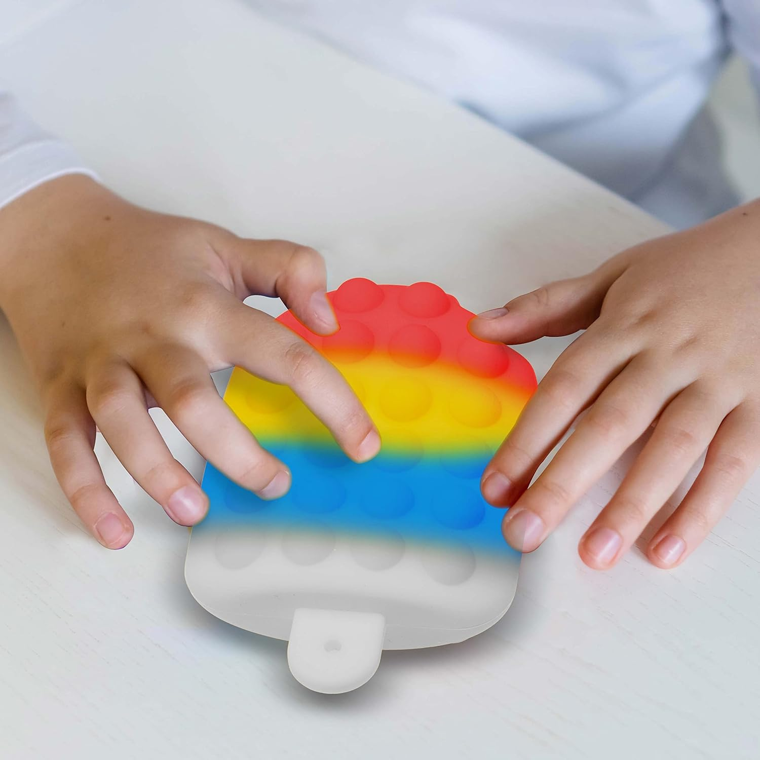 Kinderhände drücken ein buntes Pop-it-Spielzeug auf einem weißen Tisch. Das Spielzeug hat rote, gelbe und blaue Abschnitte.