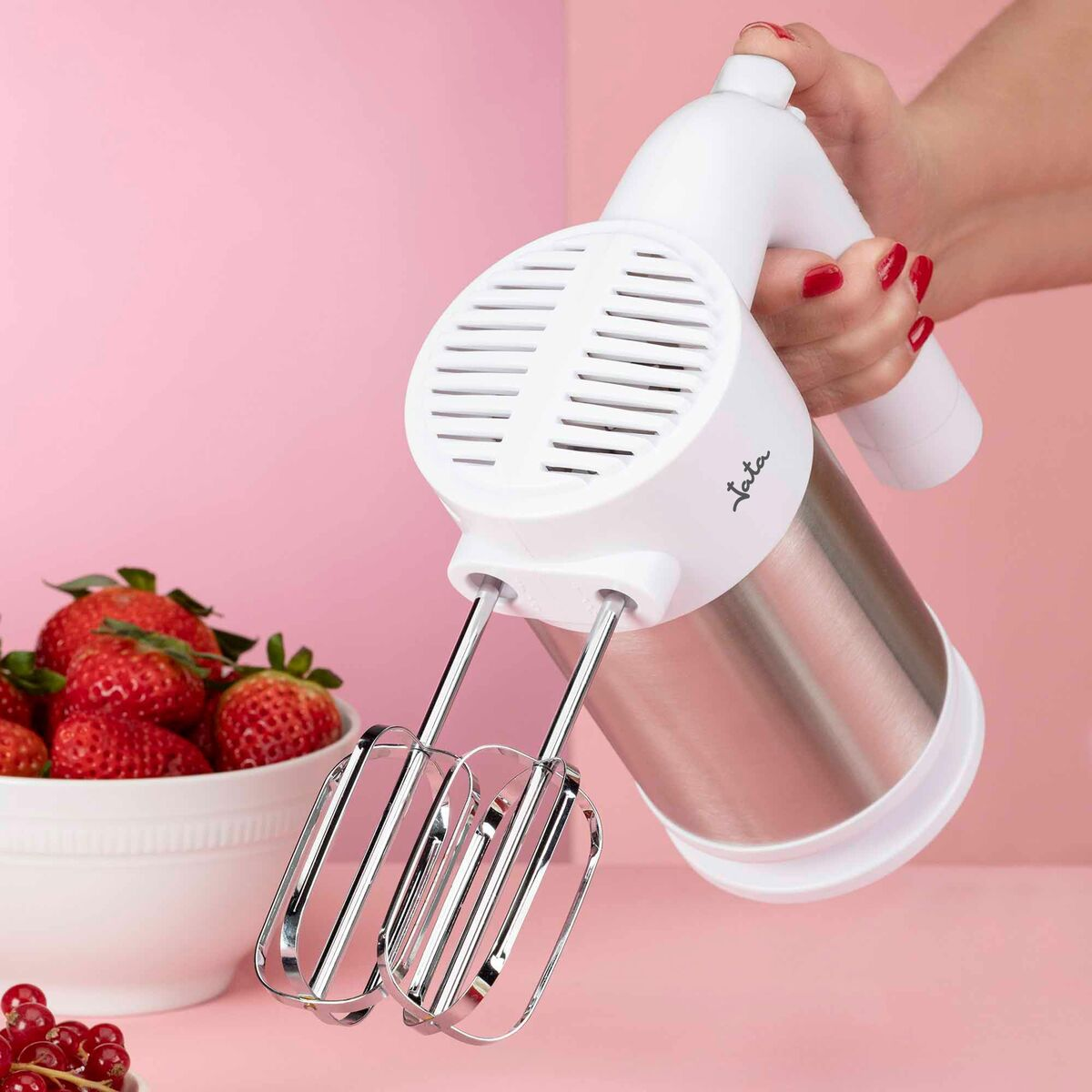 Un frullatore elettrico bianco è tenuto da una mano con smalto rosso. Fragole in una ciotola a sinistra. Lo sfondo è rosa.