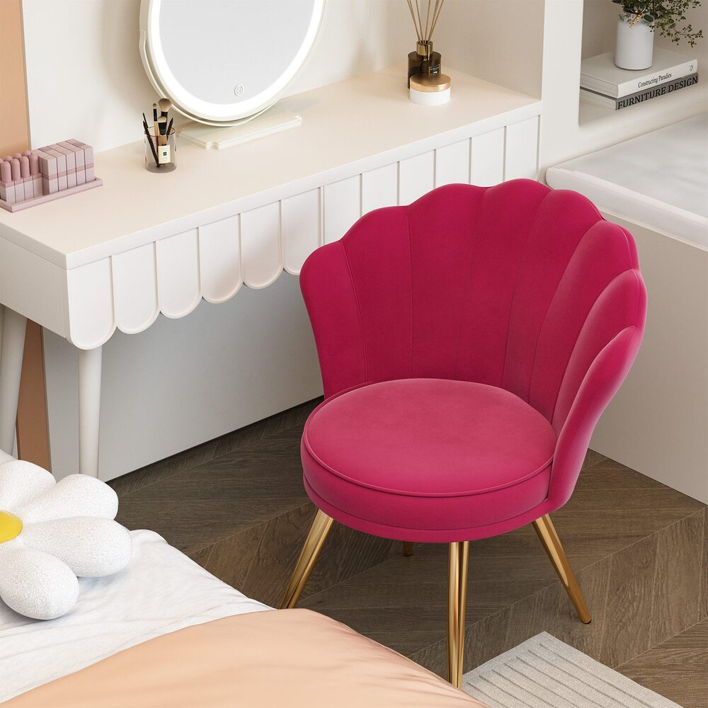 Ein pinkfarbener, muschelförmiger Stuhl mit goldenen Beinen steht neben einem weißen Schminktisch mit Spiegel. Ein Blumenkissen liegt auf dem Bett.