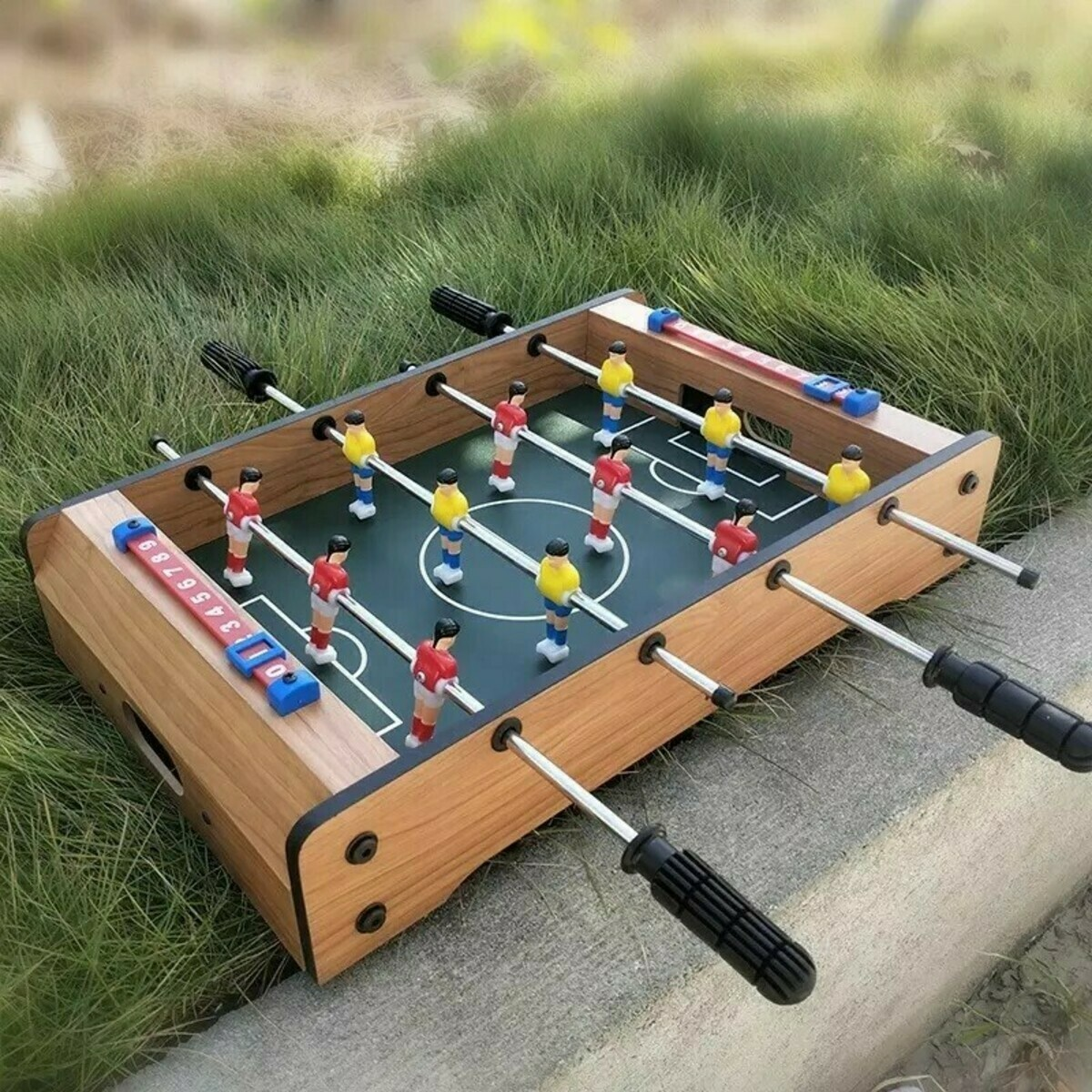Een miniatuur tafelvoetbaltafel staat op beton, omgeven door gras. Spelers in rode en gele uniformen staan op het veld.