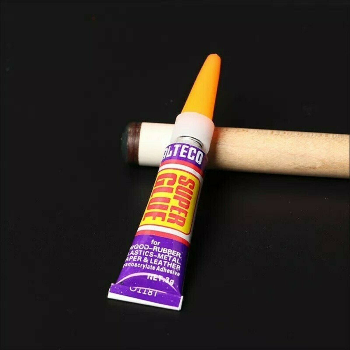 Een tube Alteco Super Glue rust op een keu. De lijmtube is paars, geel en wit. De achtergrond is een zwart oppervlak.