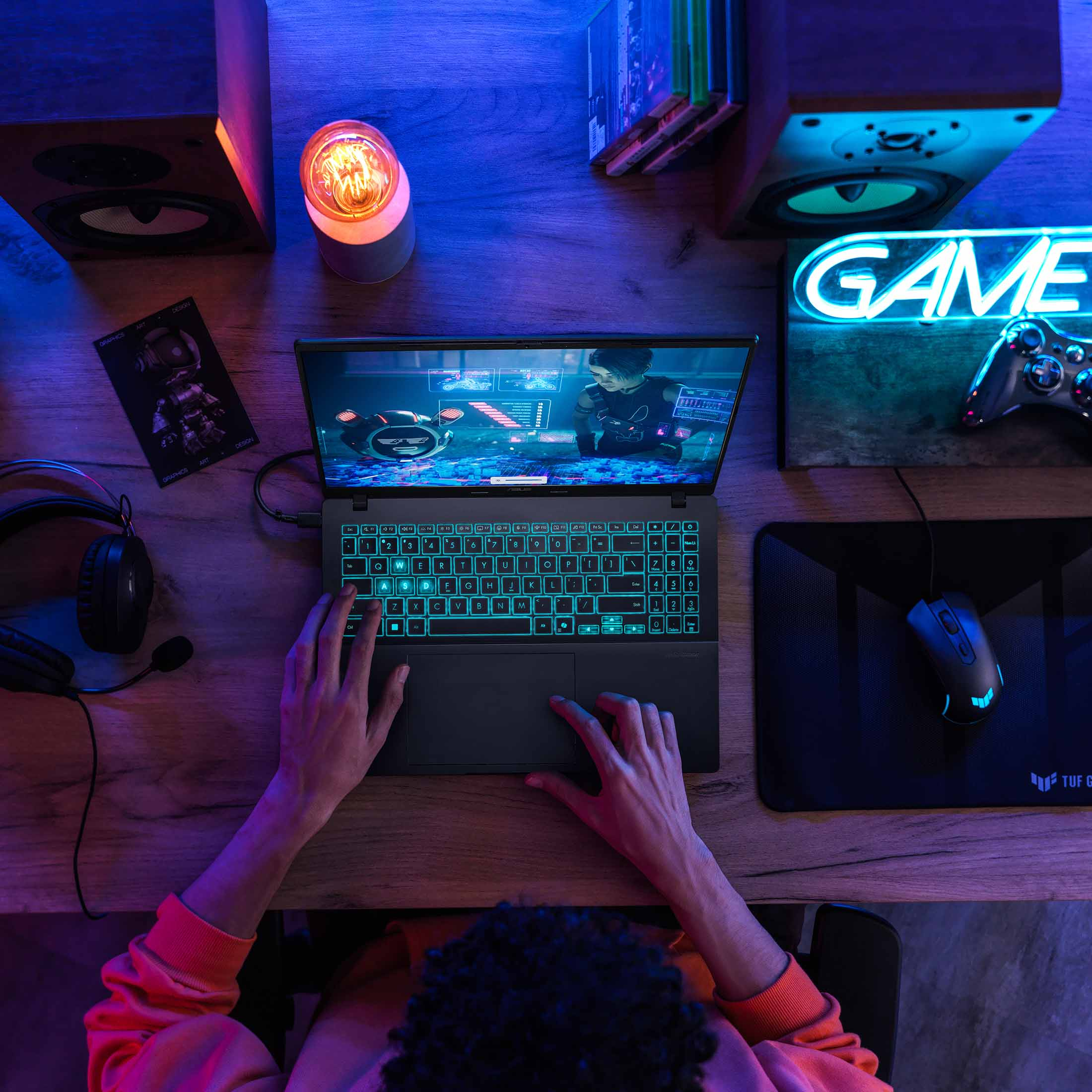 Eine Person spielt auf einem Laptop mit leuchtenden Tasten, umgeben von Lautsprechern, einer Maus und einem Neon-„GAME“-Schild.