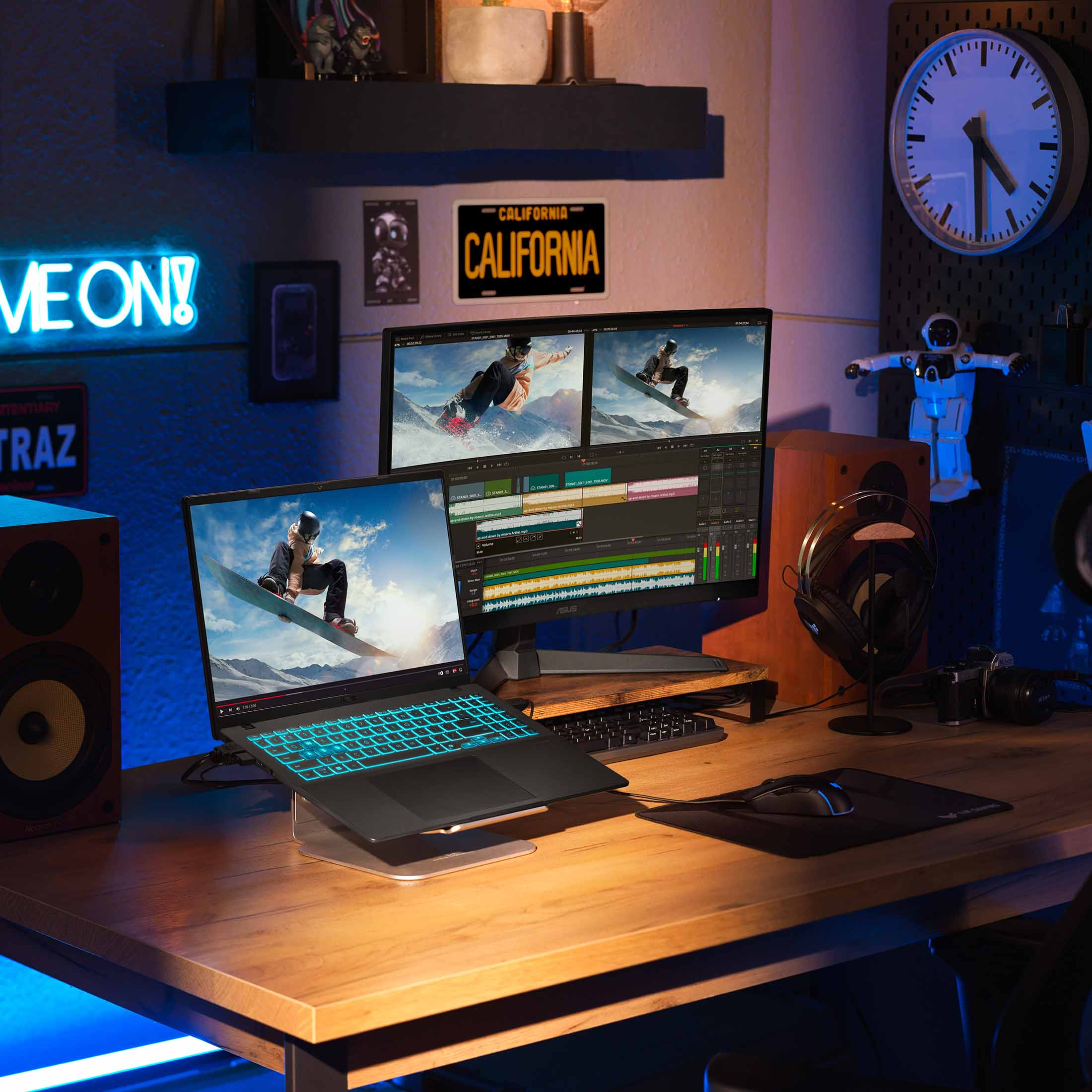 Ein Schreibtisch-Setup mit einem Laptop, mehreren Monitoren und einem Snowboarder-Bild auf dem Bildschirm. Neon-Schilder und Uhr sind sichtbar.