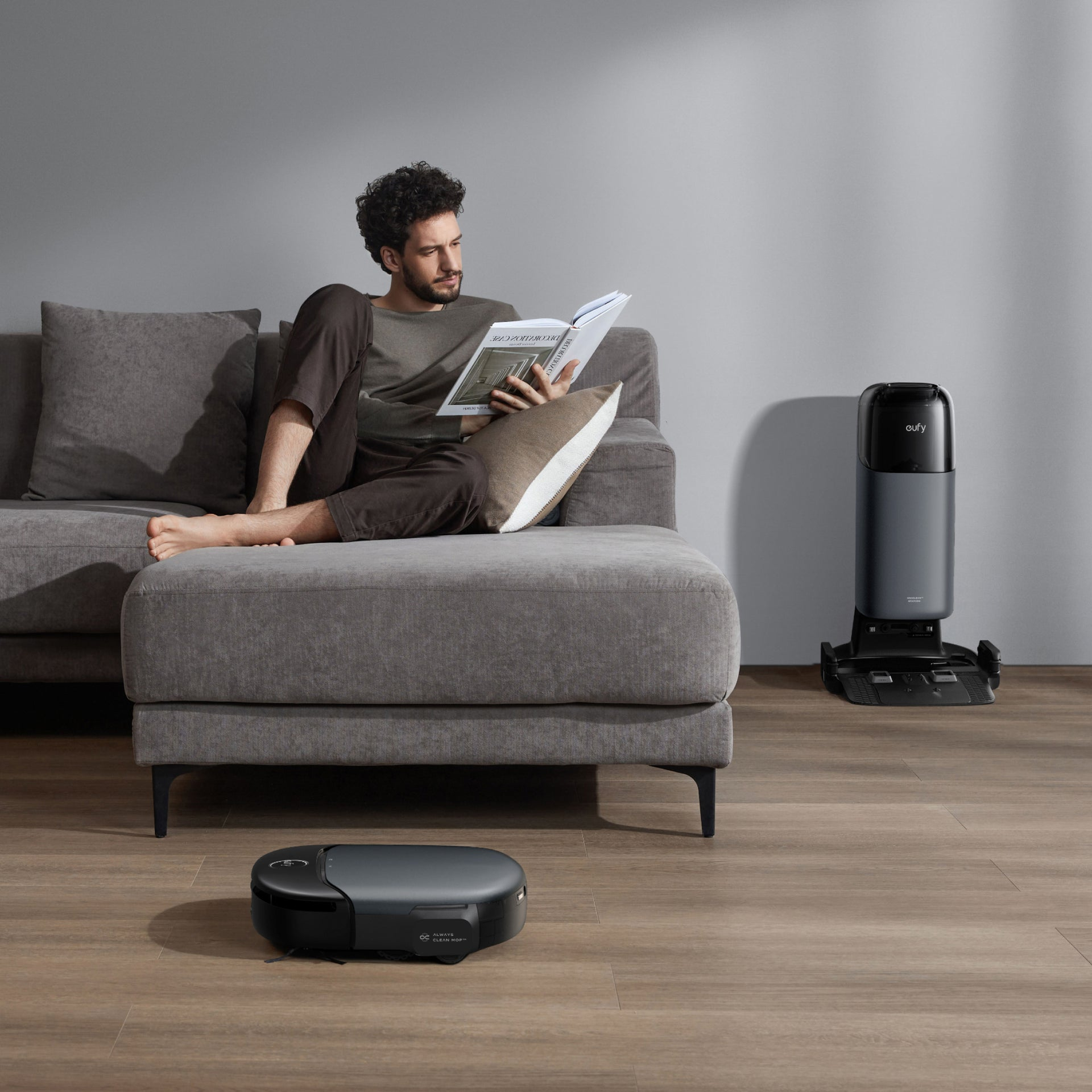 Mężczyzna czyta książkę na szarej sofie. Odkurzacz robot stoi na podłodze. Stacja czyszcząca jest po prawej.