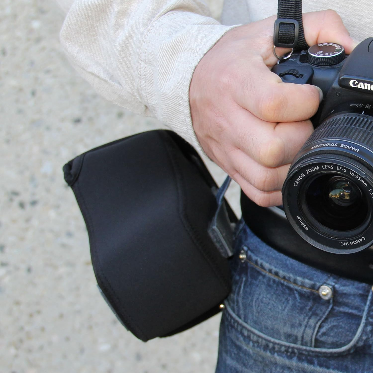 Eine Person hält eine Canon-Kamera. Die Person trägt ein beiges Hemd und blaue Jeans. Eine schwarze Objektivabdeckung ist befestigt.