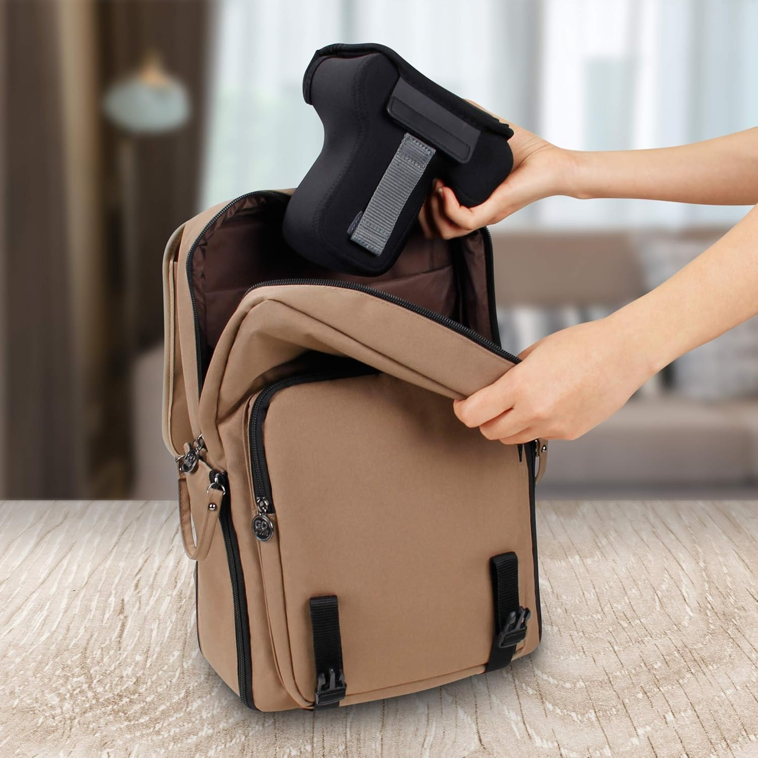 Eine Person legt eine schwarze Kameratasche in einen beigefarbenen Rucksack. Der Rucksack steht auf einer Holzoberfläche, im Hintergrund ist ein verschwommener Raum.