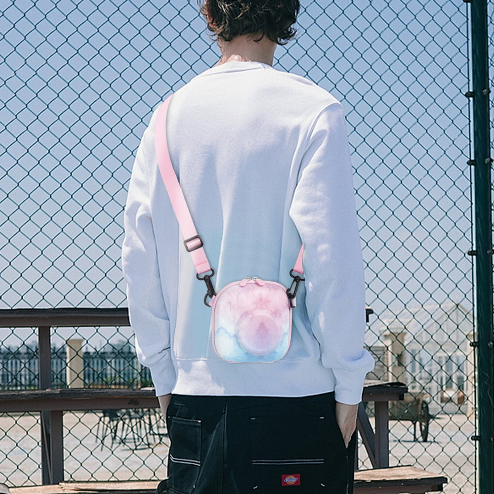 Eine Person von hinten trägt einen weißen Pullover, eine schwarze Hose und eine rosa Tasche mit buntem Muster. Im Hintergrund ist ein Maschendrahtzaun.