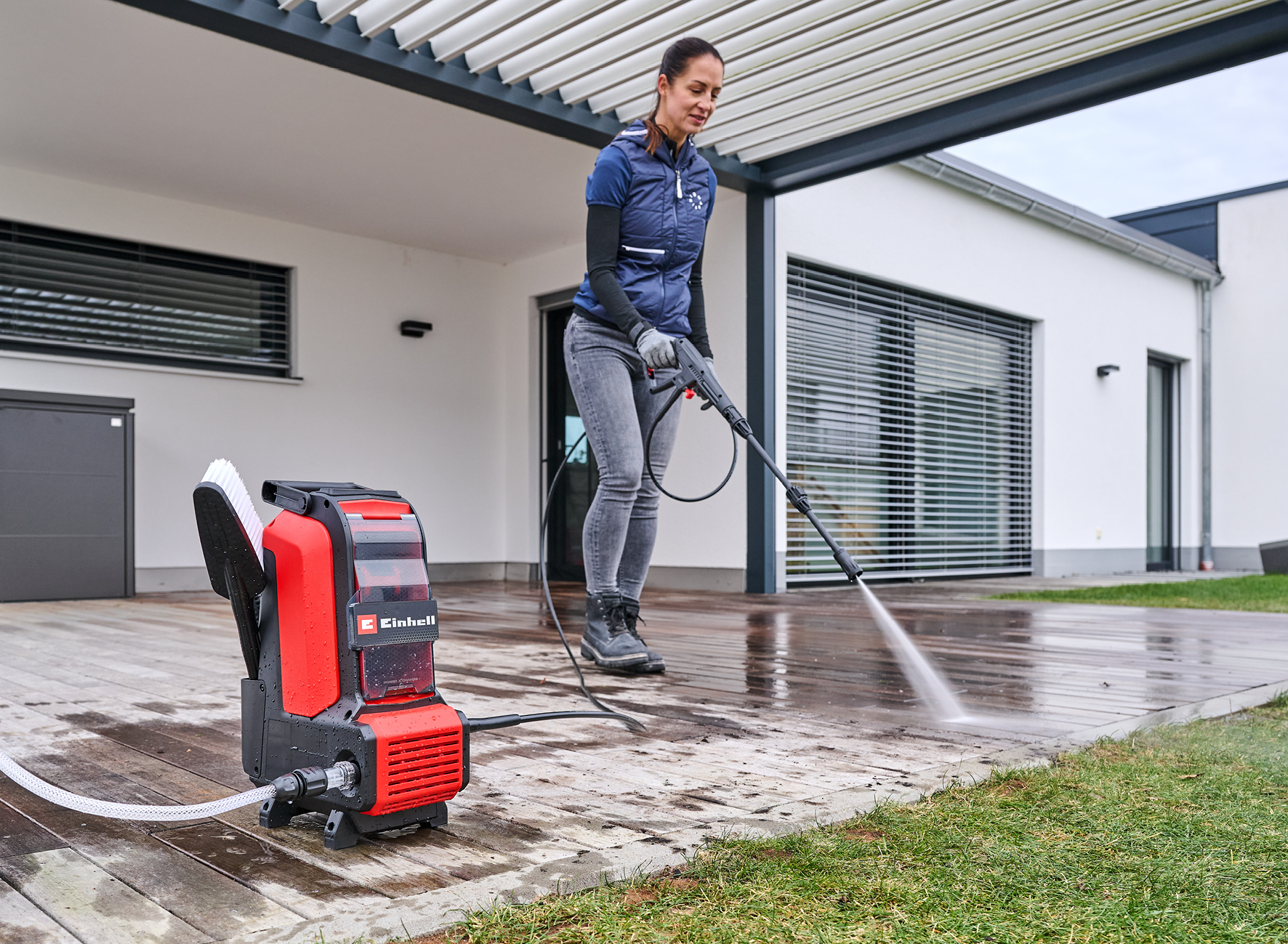 Une femme utilise un nettoyeur haute pression sur une terrasse en bois. Le nettoyeur rouge et noir est au premier plan, une maison moderne en arrière-plan.