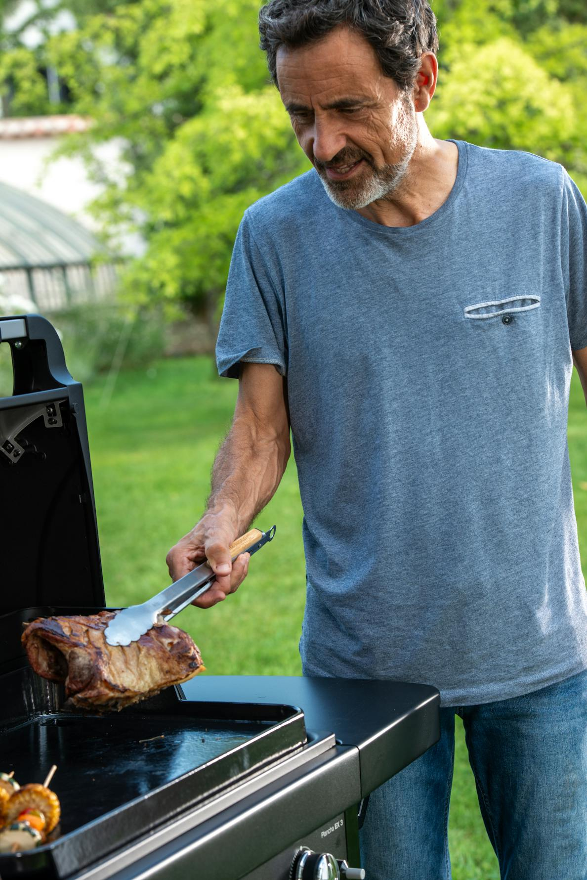 Mężczyzna grilluje mięso na zewnątrz. Używa szczypiec do obsługi dużego kawałka gotowanego mięsa na czarnym grillu.