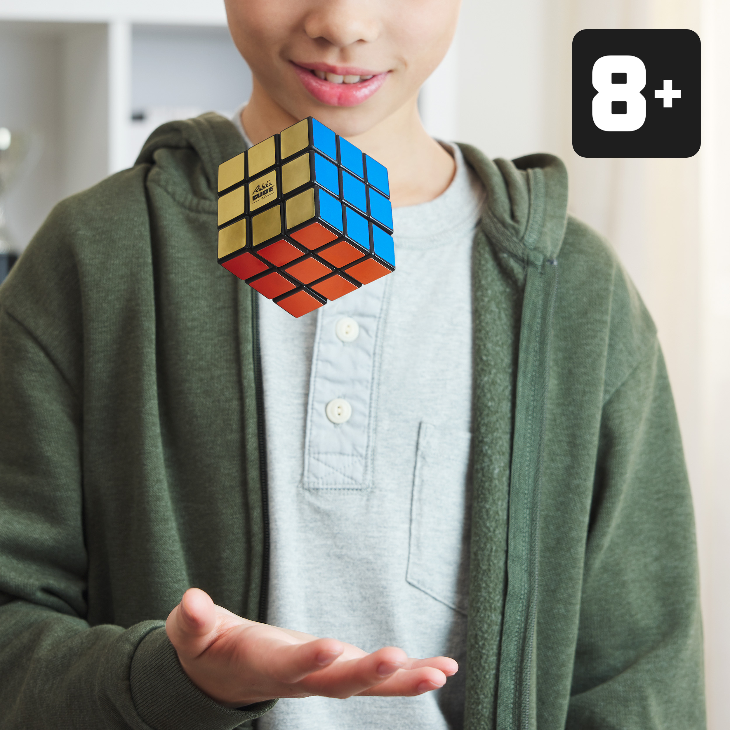 Un ragazzo sorride, tenendo un cubo di Rubik. Il cubo è sopra la sua mano. Un segno 8+ è in alto a destra.