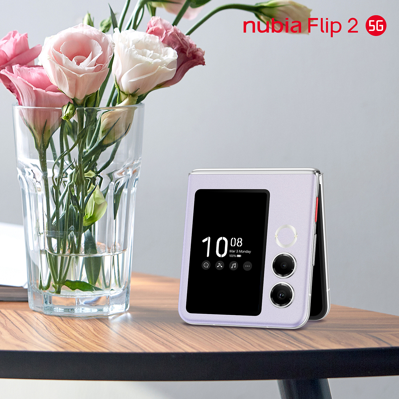 Fioletowy telefon nubia Flip 2 5G leży na drewnianym stole obok wazonu z różowymi i białymi kwiatami. Ekran wyświetla godzinę.