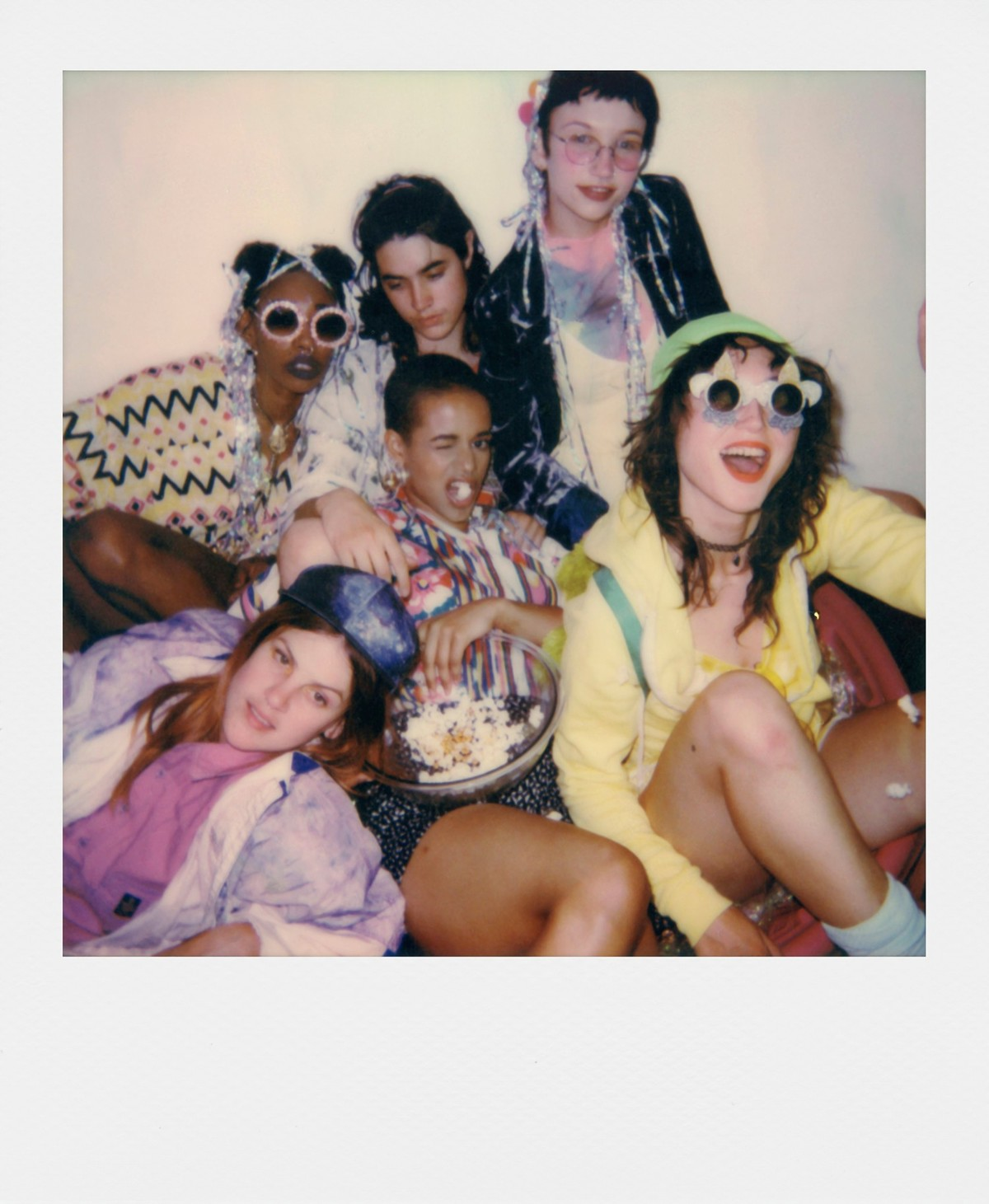 Grupo de jóvenes con gafas de sol y ropa colorida. Uno guiña un ojo. Tazón de palomitas en el medio. Foto estilo Polaroid.