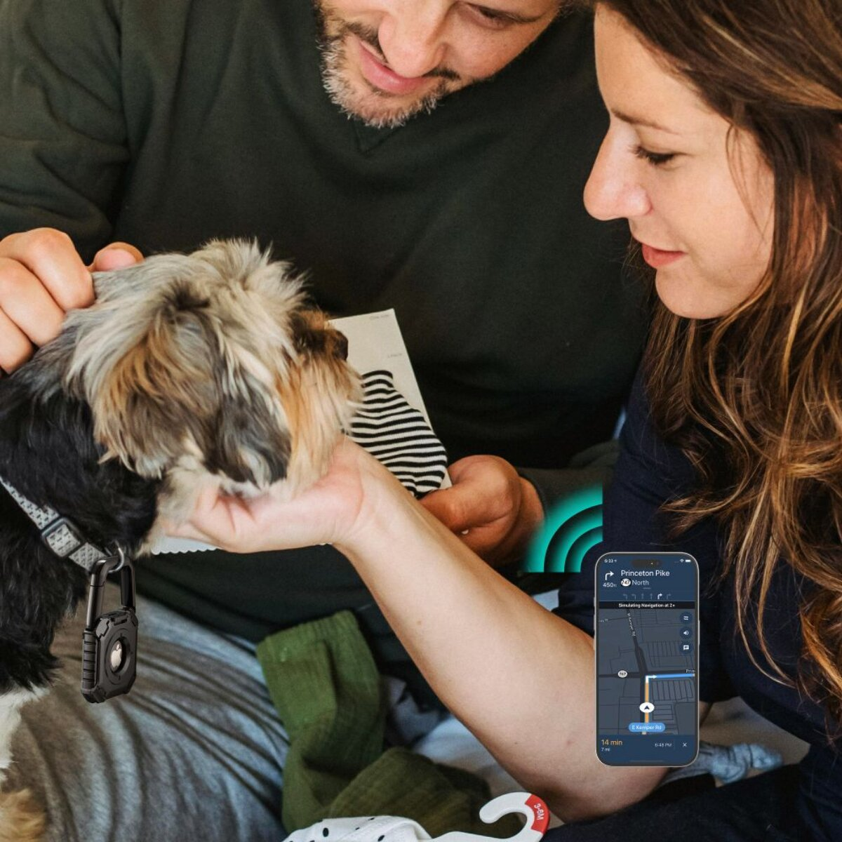 Ein Paar streichelt einen kleinen Hund. Eine GPS-Karte befindet sich auf einem Telefon. Der Hund hat ein schwarzes Ortungsgerät. Kleidung und Socken sind im Hintergrund.