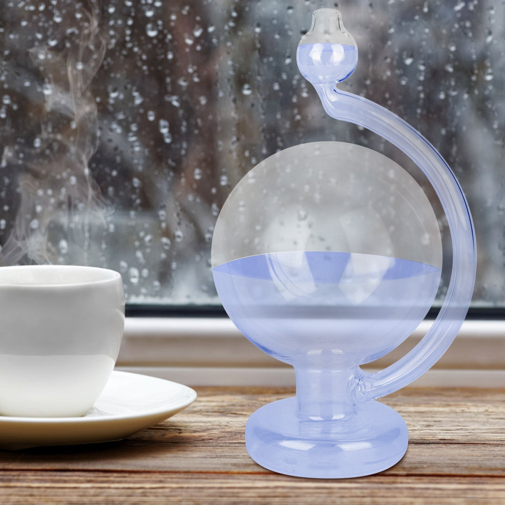 Eine weiße Tasse auf einer Untertasse, ein blaues Glaswetterinstrument und ein regnerisches Fenster stehen auf einer Holzoberfläche.