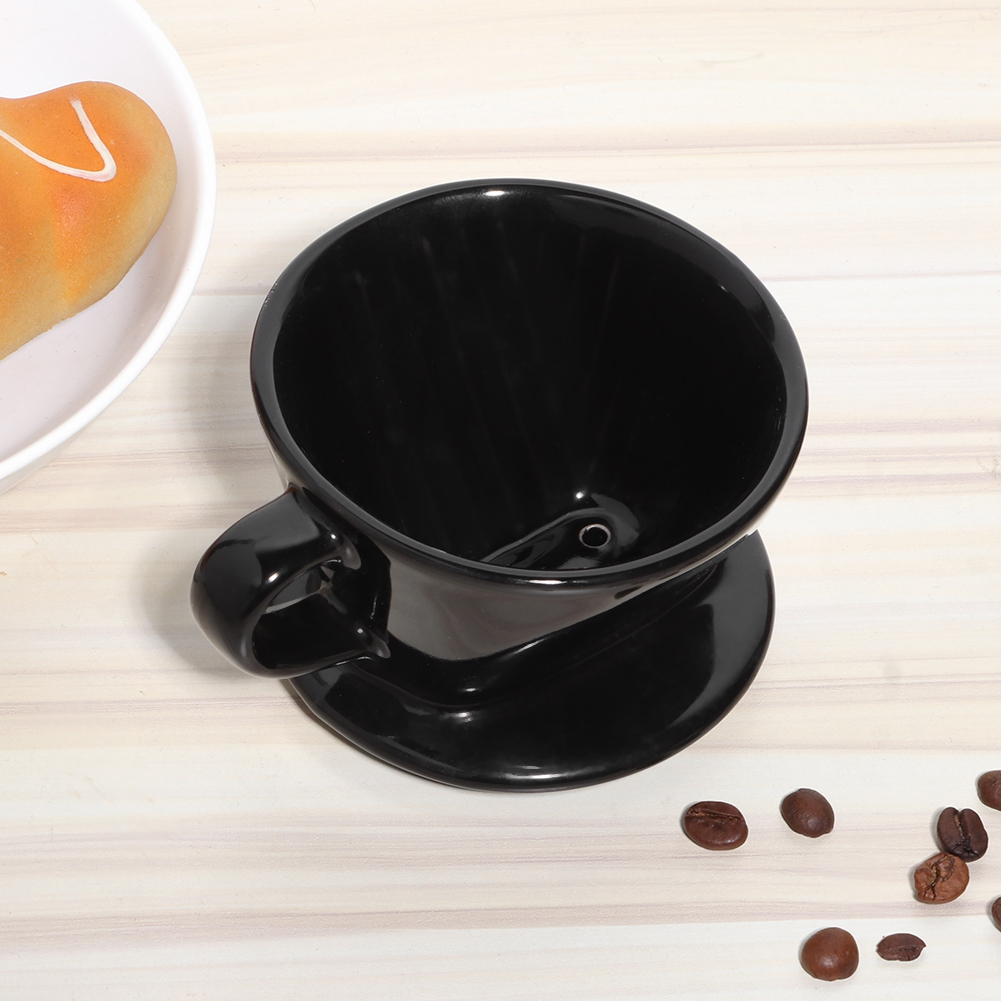 Schwarzer Kaffeetropfer mit Griff auf einer Untertasse. Kaffeebohnen sind verstreut. Ein Teller mit Gebäck ist im Hintergrund.