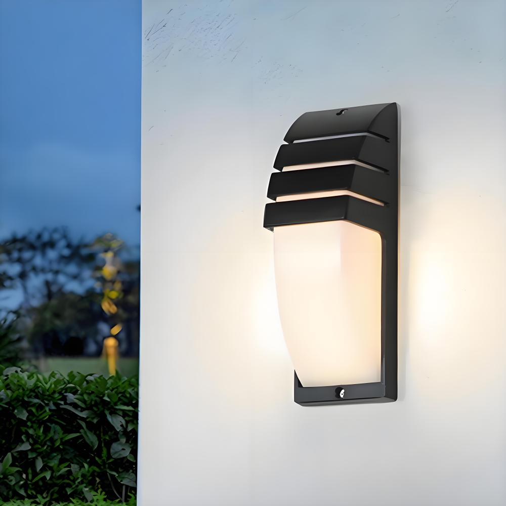 Eine schwarze Außenwandleuchte mit weißem Licht, an einer weißen Wand montiert. Der Hintergrund zeigt einen dunkelblauen Himmel und Grün.