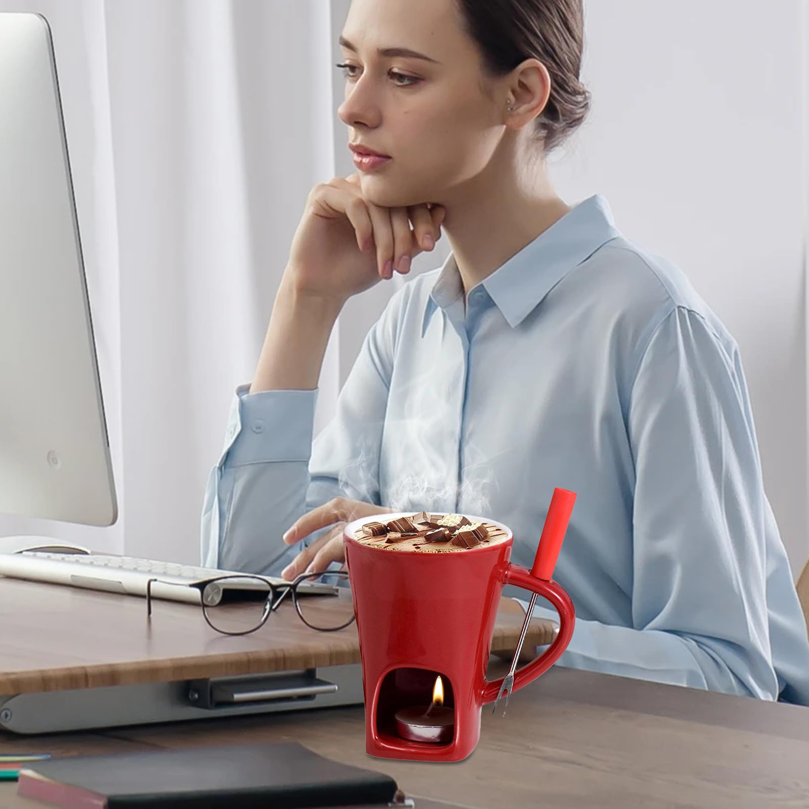 Eine Frau sitzt an einem Schreibtisch und schaut auf einen Computer. Auf dem Tisch steht eine rote Tasse mit Schokolade und eine Kerze.