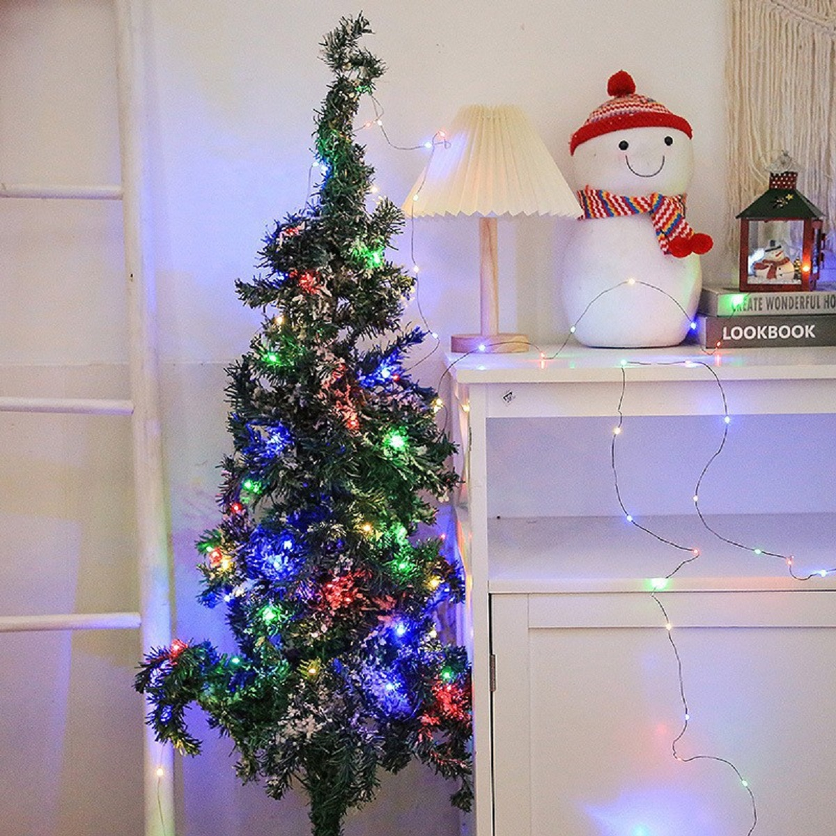 Ein kleiner Weihnachtsbaum mit bunten Lichtern steht neben einem weißen Schrank, einer Lampe und einer Schneemann-Dekoration.