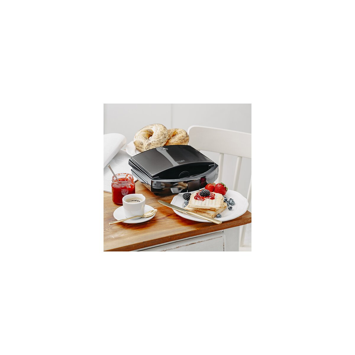 Frühstücksszene mit Waffeln, Beeren, Kaffee und Marmelade. Ein schwarzer Waffeleisen steht daneben.