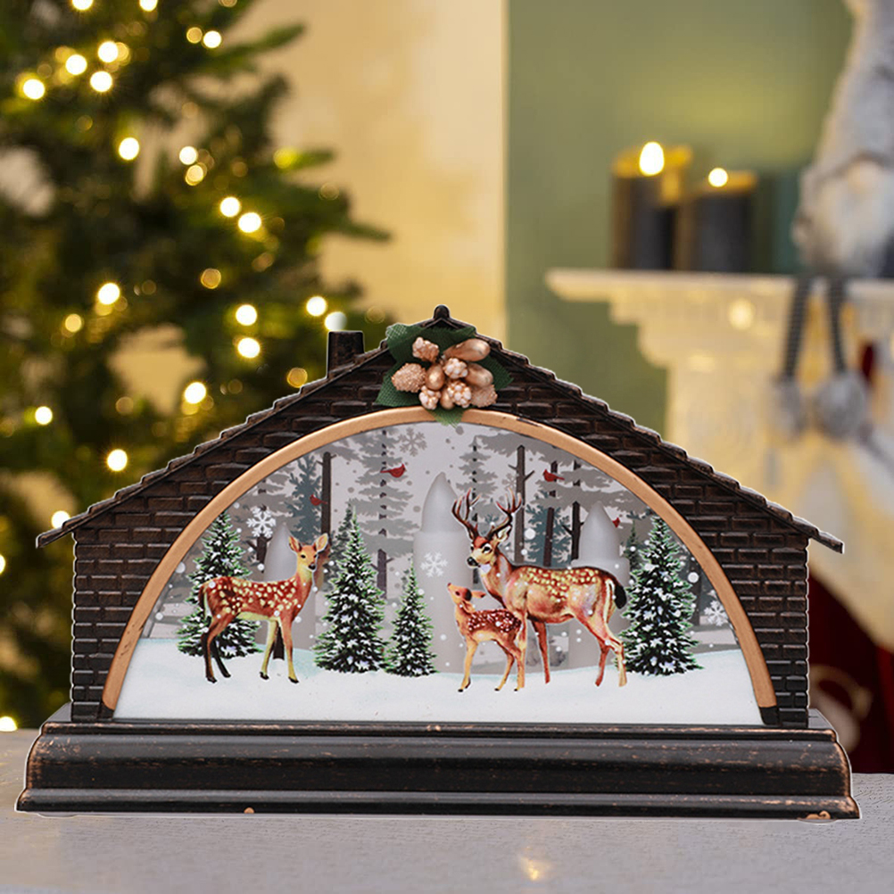 Eine dekorative Weihnachtsszene mit Hirschen in einem verschneiten Wald, in einem hausförmigen Ornament, mit einem Weihnachtsbaum im Hintergrund.
