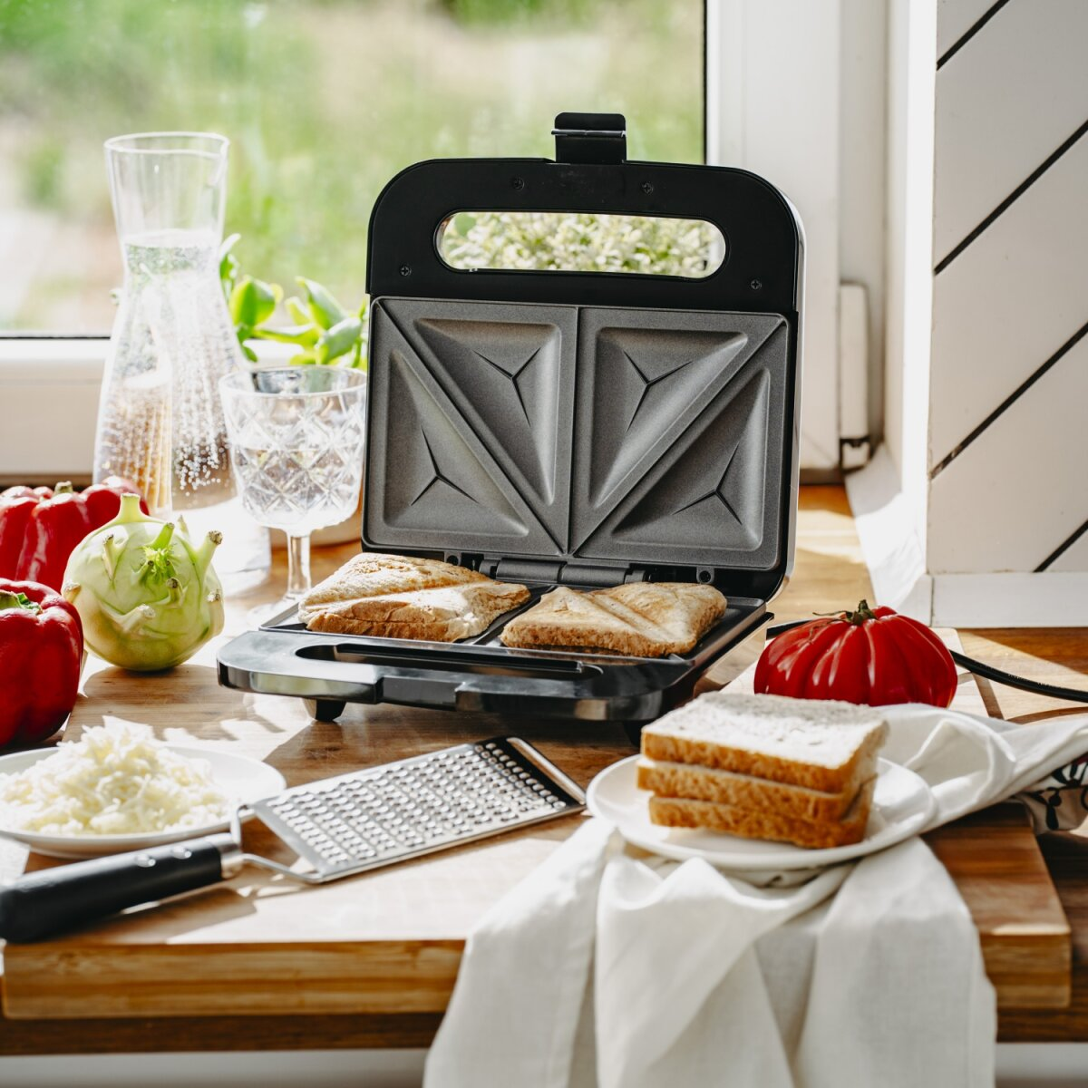 Sandwichmaker mit Sandwiches, Brot, Käse, Gemüse und einer Reibe auf einem Holztisch, in der Nähe eines Fensters.