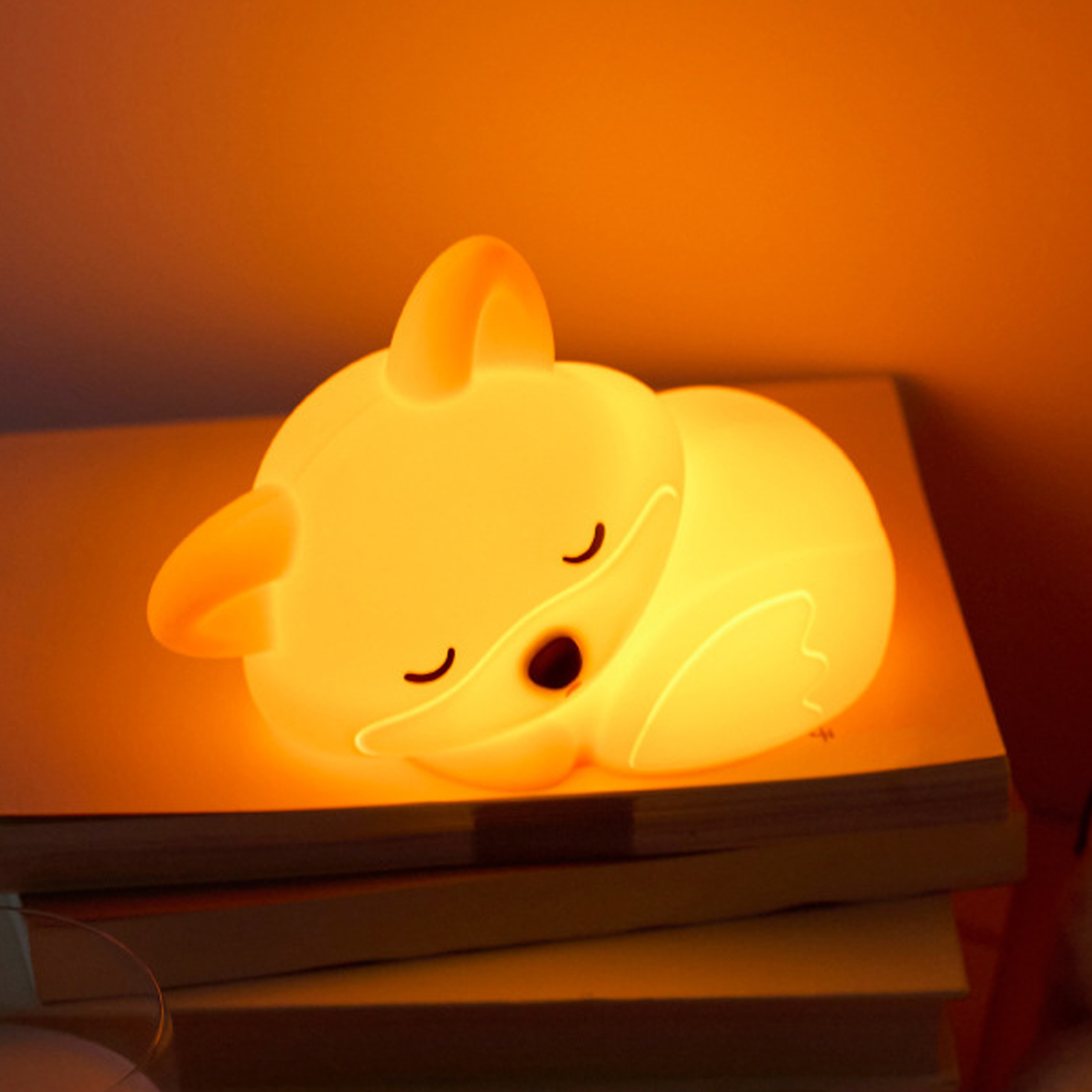 Ein leuchtendes Fuchsnachtlicht liegt auf Büchern. Der Fuchs ist weiß und orange, mit geschlossenen Augen. Der Hintergrund ist orange.