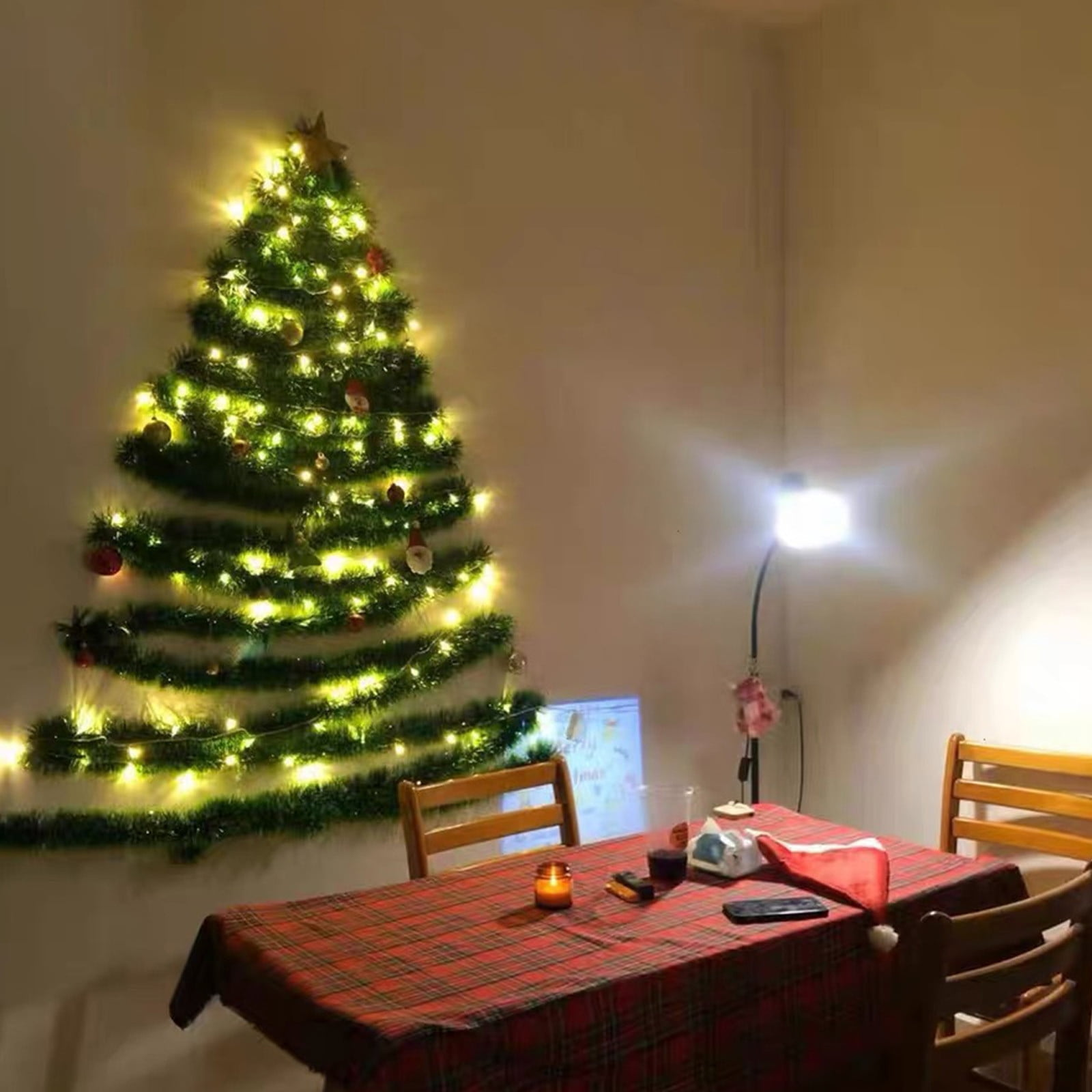 Ein Weihnachtsbaum aus Girlanden an einer Wand, mit Lichtern und Ornamenten. Davor ein gedeckter Tisch.