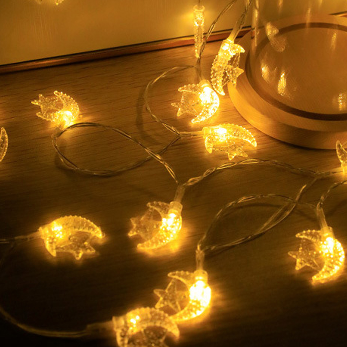 Lichterketten mit Mond- und Sternformen beleuchten eine Holzoberfläche. Ein Glasbehälter steht im Hintergrund.