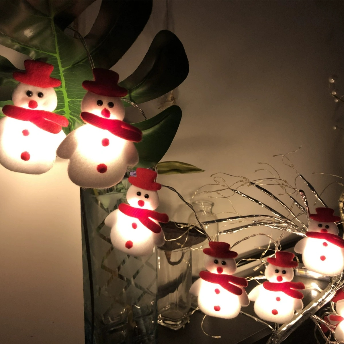 Lichterketten mit Schneemann-Ornamenten. Jeder Schneemann hat einen roten Hut und Schal, mit leuchtendem Körper. Sie sind in der Nähe von grünen Blättern.