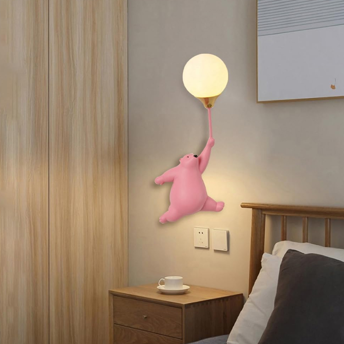 Eine rosa bärenförmige Lampe hält eine ballonförmige Leuchte. Sie ist an einer grauen Wand befestigt. Darunter befinden sich ein Nachttisch und ein Bett.
