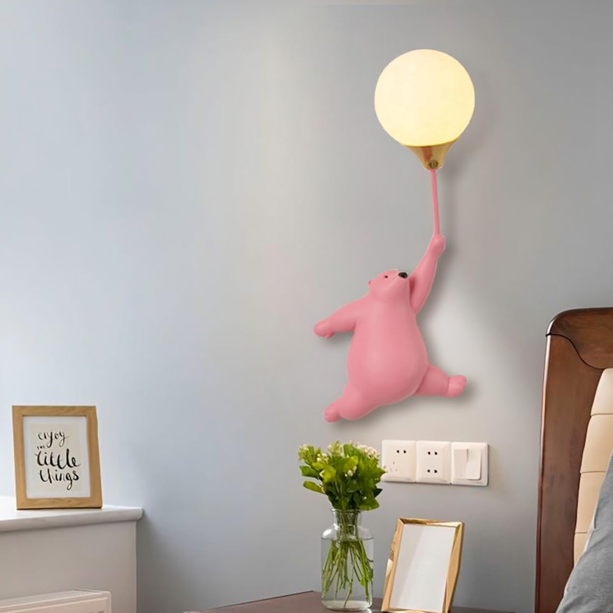 Eine rosa bärenförmige Lampe hält eine Ballonlampe. Die Lampe befindet sich an einer grauen Wand. Darunter ein Bilderrahmen und Blumen.