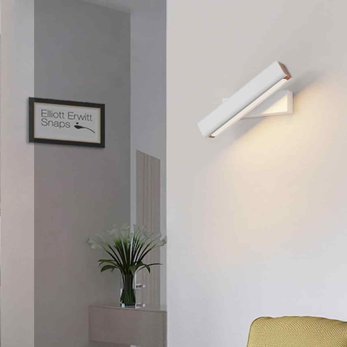 Eine moderne Wandlampe ist an einer weißen Wand angewinkelt. Ein gerahmtes Kunstwerk und eine Blumenvase sind sichtbar. Ein gelbes Sofa ist in der Ecke.