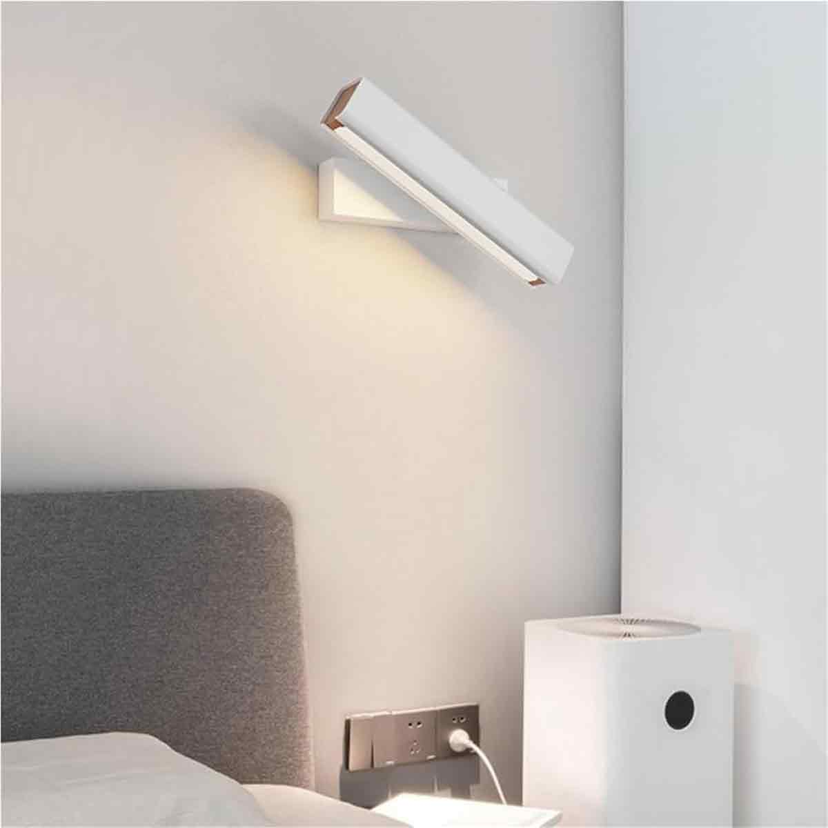 Eine moderne weiße Wandleuchte beleuchtet ein Schlafzimmer. Ein graues Kopfteil ist sichtbar, zusammen mit einem Luftreiniger und einer Steckdose.